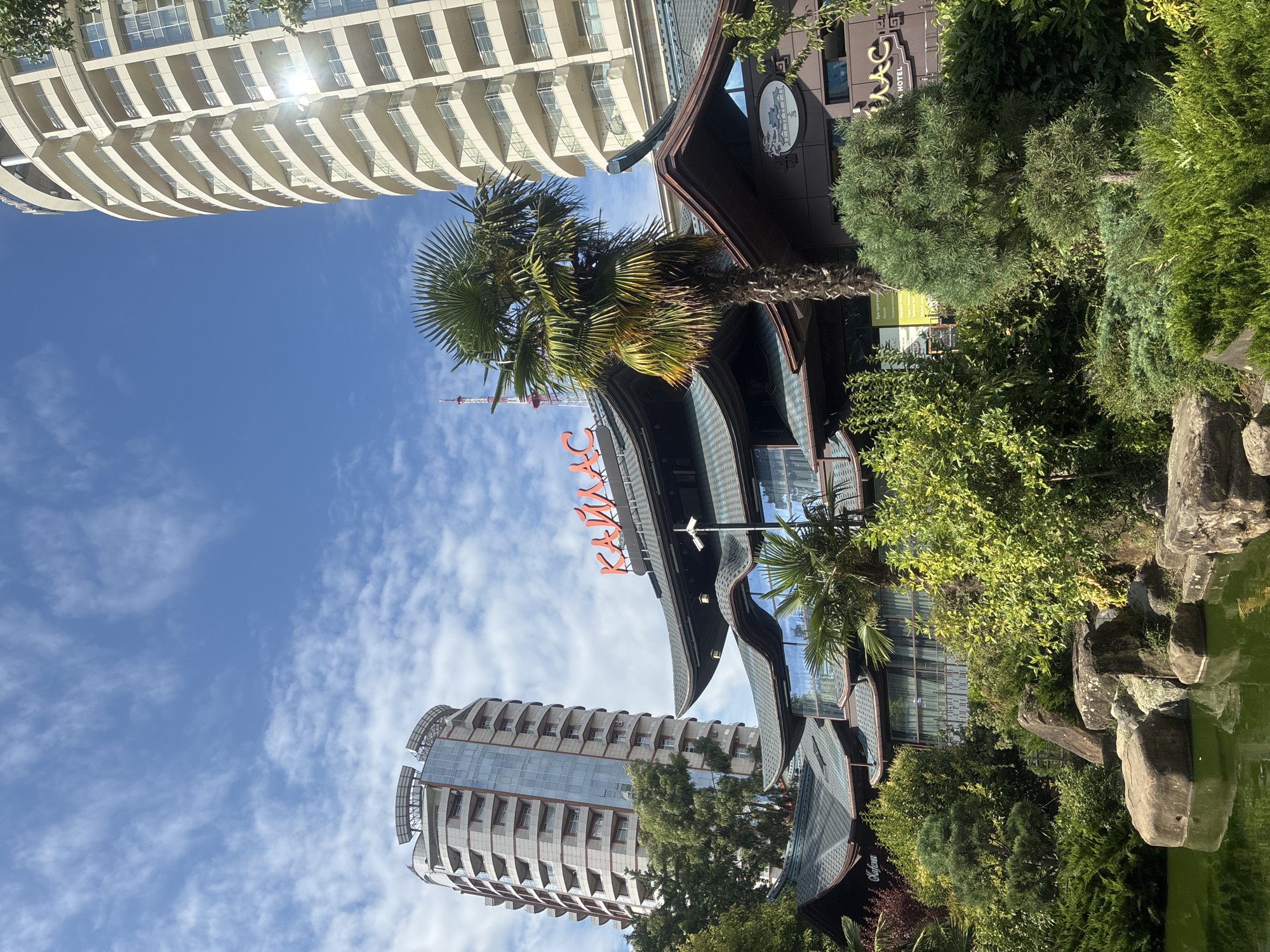 The image shows a view of a building complex with traditional East Asian architectural elements, including curved roofs and ornate details. The building is primarily dark brown with teal accents on the roof. A large red sign with Cyrillic lettering, "КАЙЛАС," is prominently displayed on the front of the building. The building is surrounded by lush greenery, including palm trees and various shrubs, and a small pond with large rocks in the foreground. In the background, two modern high-rise apartment buildings are visible, one on the left and another taller one on the right. The sky is blue with scattered white clouds. The building with the "КАЙЛАС" sign appears to be a hotel, as indicated by the word "HOTEL" below the name. The location is identified as Sochi, Russia. There are no people or discernible actions depicted in the image.