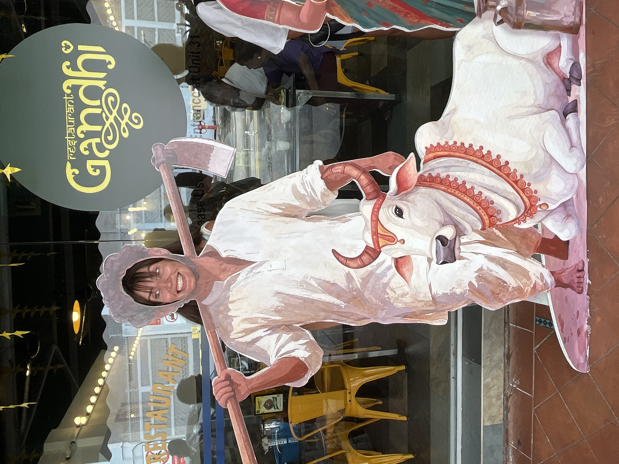 This is a photograph of a life-sized cardboard cutout depicting a person with a woman's face inserted into the head opening, dressed in traditional Indian farmer attire and holding a hoe. Beside the cutout is a depiction of a white cow lying down, adorned with a red and gold necklace. The cutout is situated outside the "Restaurant Gandhi" in Singapore, as indicated by the prominent signage. In the background, through the restaurant window, several patrons are visible dining at tables, with yellow chairs and restaurant decor also present. The overall setting is a street-facing storefront.