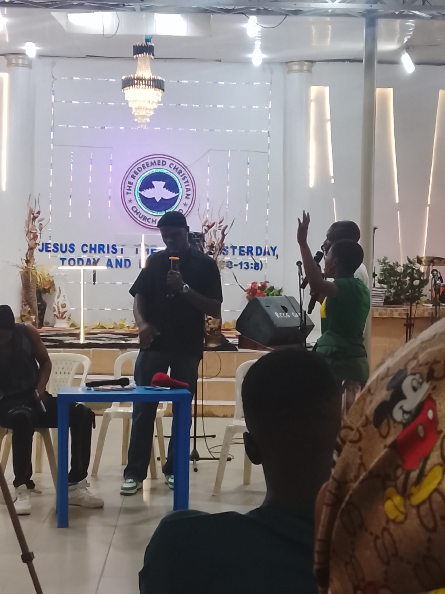 The image depicts a church setting, possibly The Redeemed Christian Church, with a stage in the background displaying the church's logo and scripture. A man in dark clothing stands at a blue table, holding a microphone and appearing to address the audience. To his right, two individuals stand with one raising their hands, seemingly engaging in worship or prayer. In the foreground, people are seated, listening to the speaker. The environment is indoors, with a focus on the stage area. There is no indication of the city or specific location within Unknown, Unknown.