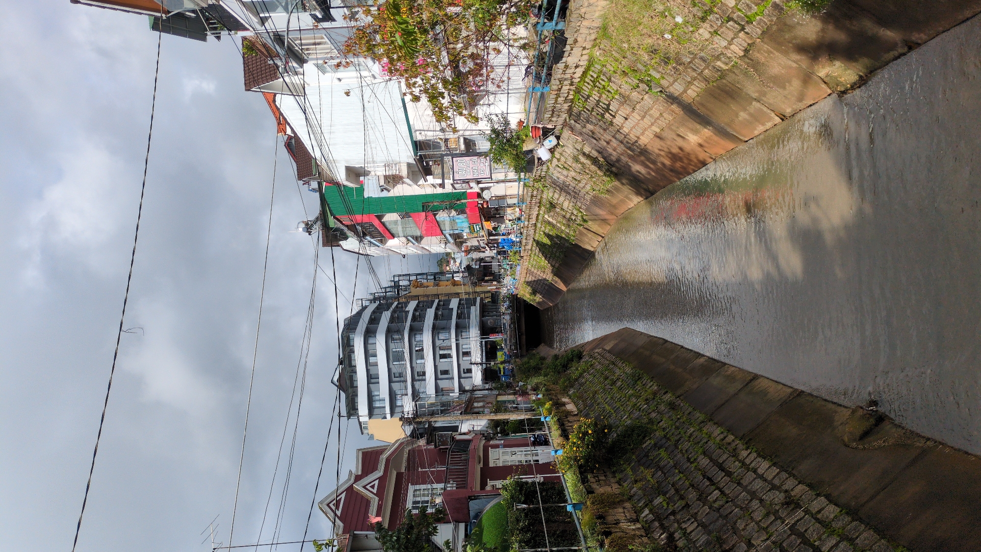 The image depicts a view of a canal in Da Lat, Vietnam. The canal has concrete-lined sides with stone-like brickwork on the upper portions, and the water within is murky brown, reflecting the overcast sky and surrounding buildings. Along the canal's edges, there are grassy patches and some scattered vegetation.
On either side of the canal, urban buildings are visible. On the left, a distinctive red building with white trim and peaked roofs stands out, with a modern, curved white building behind it. The right side of the canal features a row of multi-story buildings painted in various colors, including white, green, and red. Some of these buildings have balconies and prominent windows. A tree with pink flowers is also visible on the right.
Overhead, numerous power lines crisscross the sky, adding to the urban feel of the scene. In the distance, a bridge spans the canal, with more buildings and hills visible beyond it. There are no prominent individuals or specific actions captured in the image. The overall impression is one of a functional, urban waterway integrated into the cityscape of Da Lat.