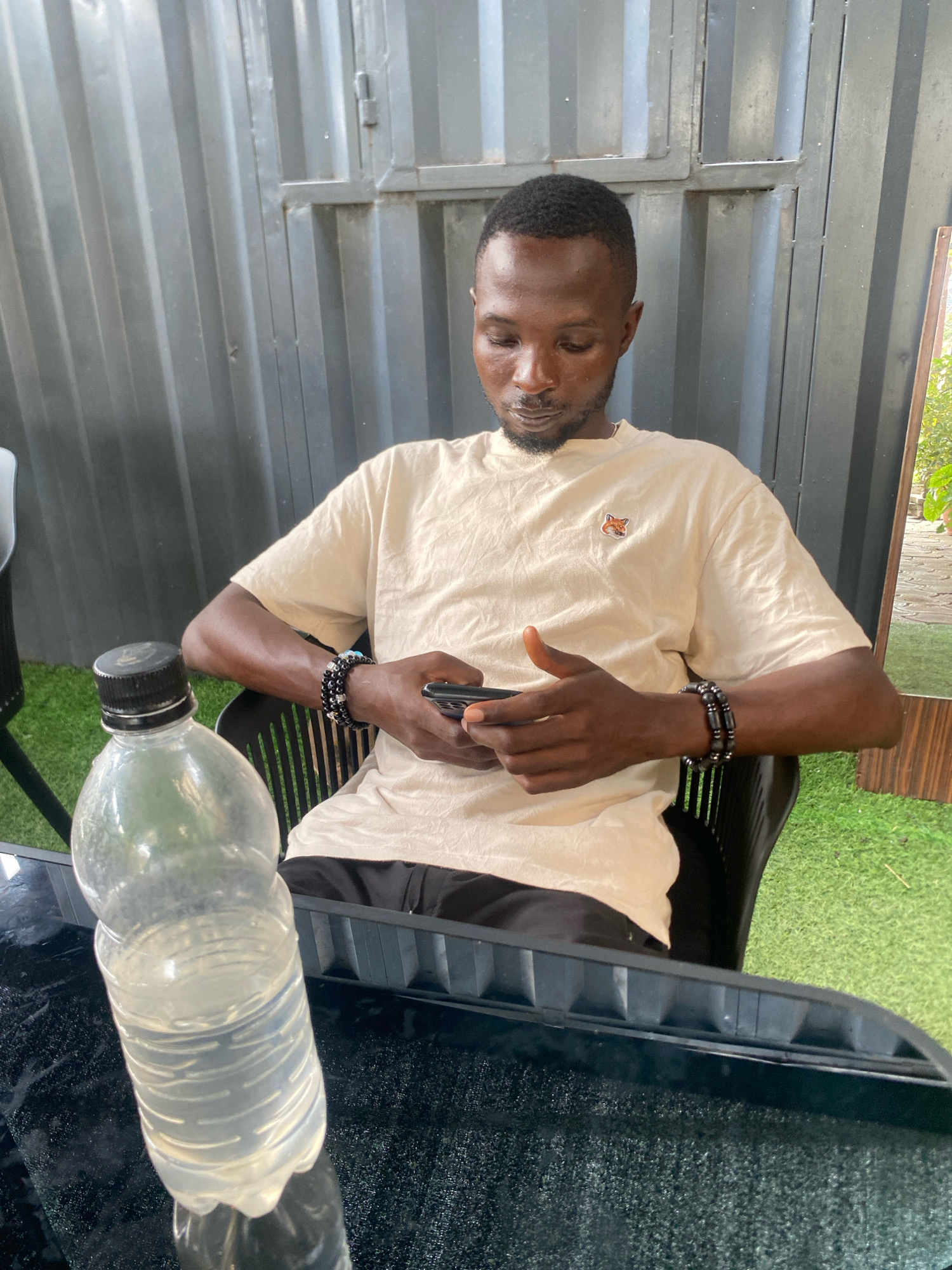 In Barden Yana, Nigeria, a young man with short black hair and a beard sits in a black plastic chair. He is wearing a light beige t-shirt with a small embroidered fox logo on the left chest. He has two beaded bracelets on his left wrist, one black with spherical beads and the other with faceted black beads. He is looking down at his phone, which he holds in both hands. In the foreground, a clear plastic bottle filled with a light-colored liquid sits on a dark, textured surface, likely a table. The background consists of corrugated metal walls and artificial turf. A reflection of a leafy green plant is visible in the upper right corner.
