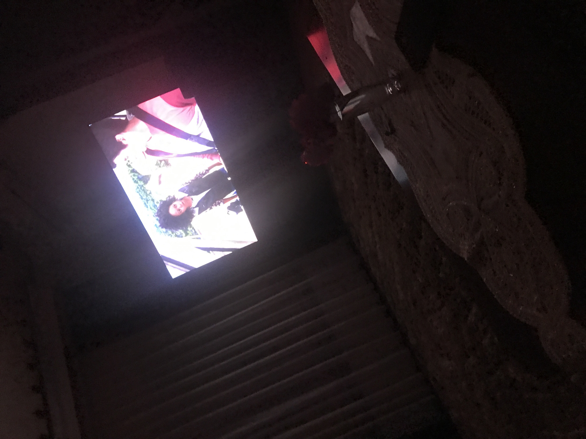 The image depicts an indoor scene in Yerevan, Armenia. A screen displays an image of two people, one with curly hair and wearing a jacket, the other with a pink item. A cigarette is visible near the screen. Adjacent to the screen is a rough, textured wall. In the foreground, the pattern of steps is seen, suggesting the photo was taken at the bottom of some steps, looking up at the screen. The ambient light is dim.