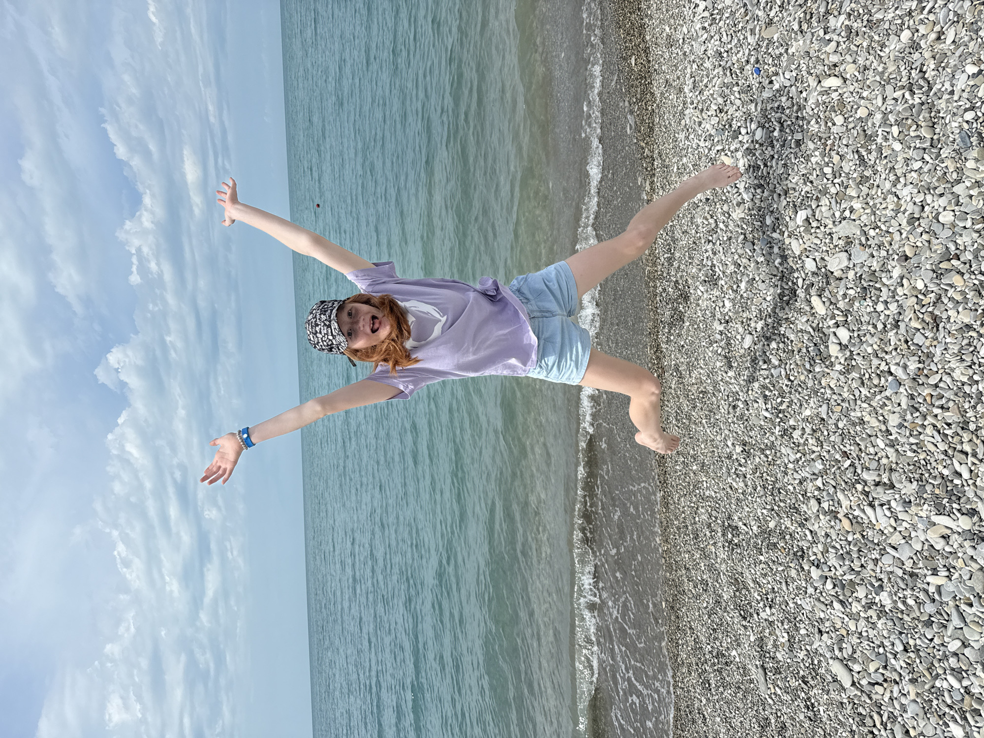 A young girl with red hair is joyfully leaping on a pebble beach with the sea and a cloudy sky in the background. She is wearing a light purple t-shirt, light blue shorts, and a black and white patterned baseball cap. Her arms are outstretched, and her legs are spread wide in a star-like pose. Her mouth is open, and she appears to be laughing or shouting. She is also wearing a blue bracelet on her left wrist. The sea is a calm, turquoise color with small waves lapping at the shore. The beach is covered in smooth, grey pebbles. The sky is a pale blue with scattered white clouds. The sun is shining, suggesting it is daytime, and the weather appears to be pleasant. The image was taken in Nebug, Russia. There is no visible text in the image.