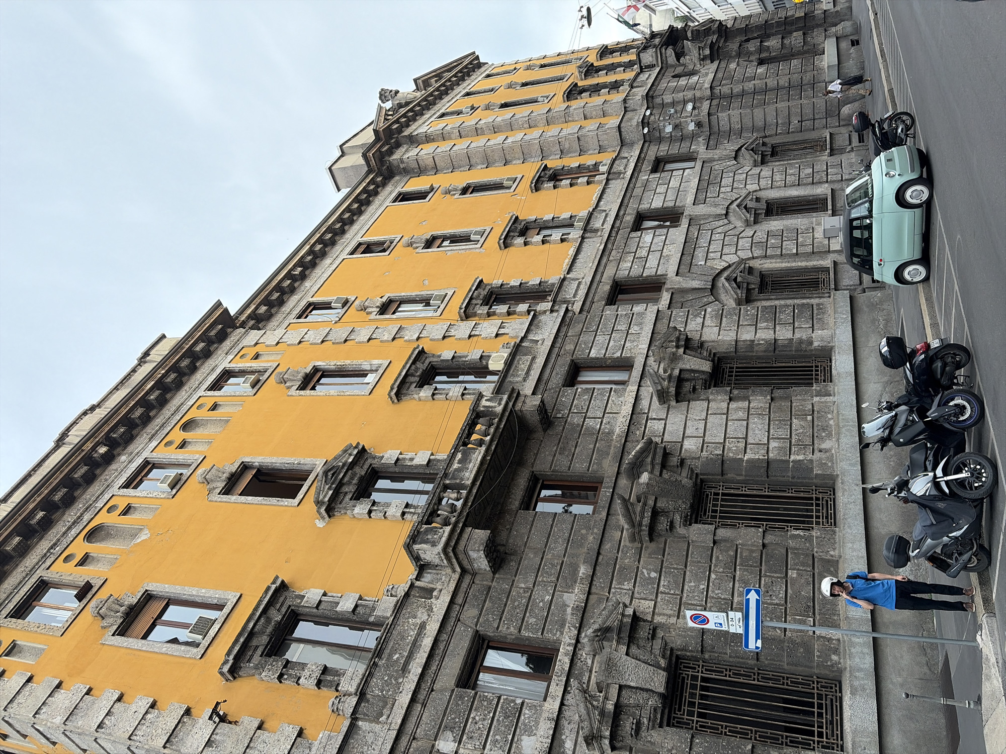 A man wearing a blue shirt, black pants, and a white helmet stands on a sidewalk in Milan, Italy, smoking a cigarette. He is standing next to a parked scooter and a light blue Fiat 500. The man is positioned in front of a large, old building with a yellow facade and stone detailing. The building has numerous windows with ornate stonework around them. Several other parked scooters and a light blue car are visible along the street. The sky is overcast.