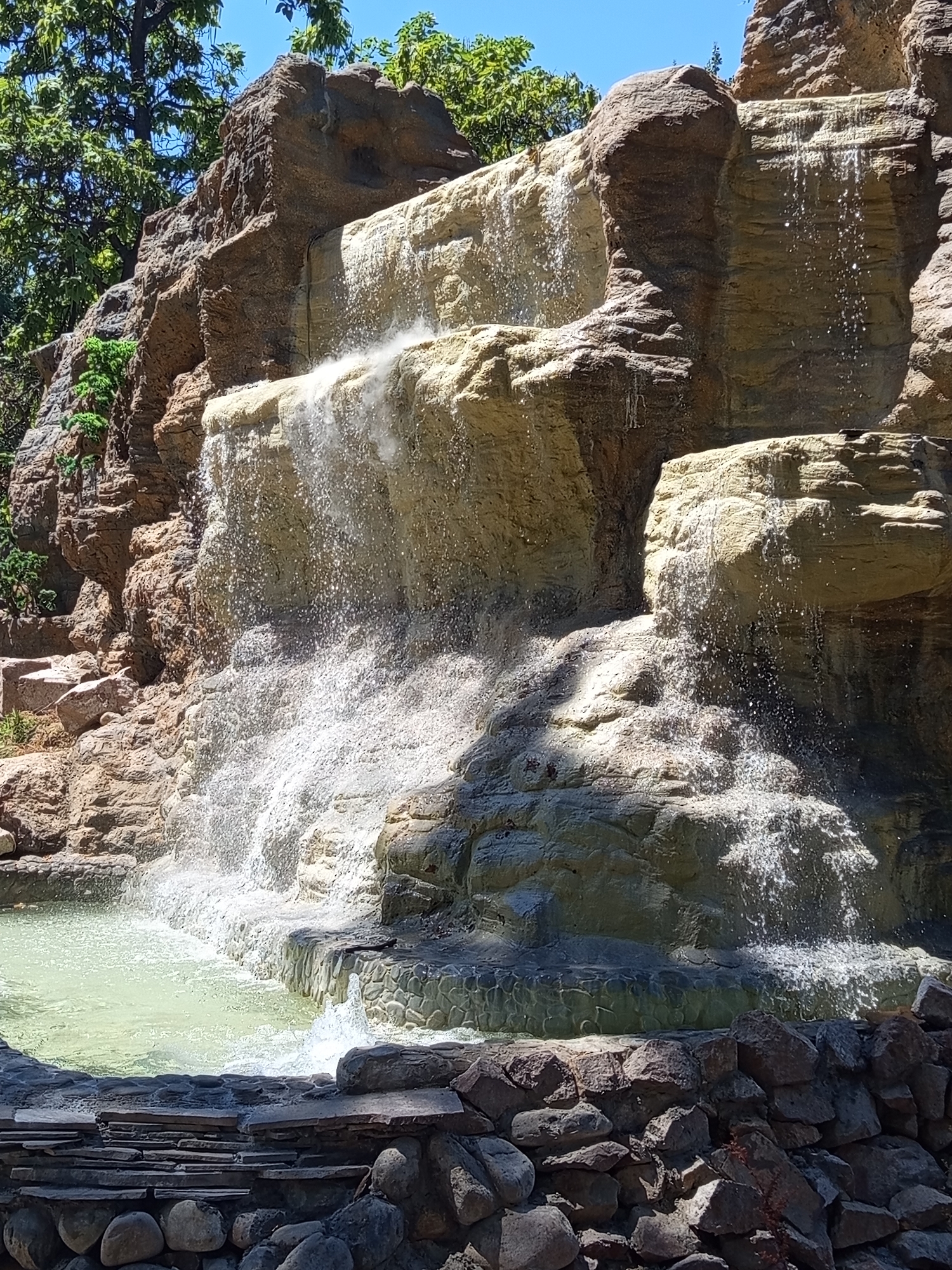 The image depicts a man-made waterfall structure in the city of Shymkent, Kazakhstan. The waterfall is integrated into a rocky, multi-tiered landscape with a pool of light-green water at its base. The rocks appear to be artificial, textured and colored to resemble natural stone. Water cascades down from multiple levels, creating a dynamic and somewhat frothy effect. Green foliage, including trees, is visible in the upper left portion of the image, suggesting an outdoor setting. The overall scene conveys a recreational or park-like environment. No people are present in the image.