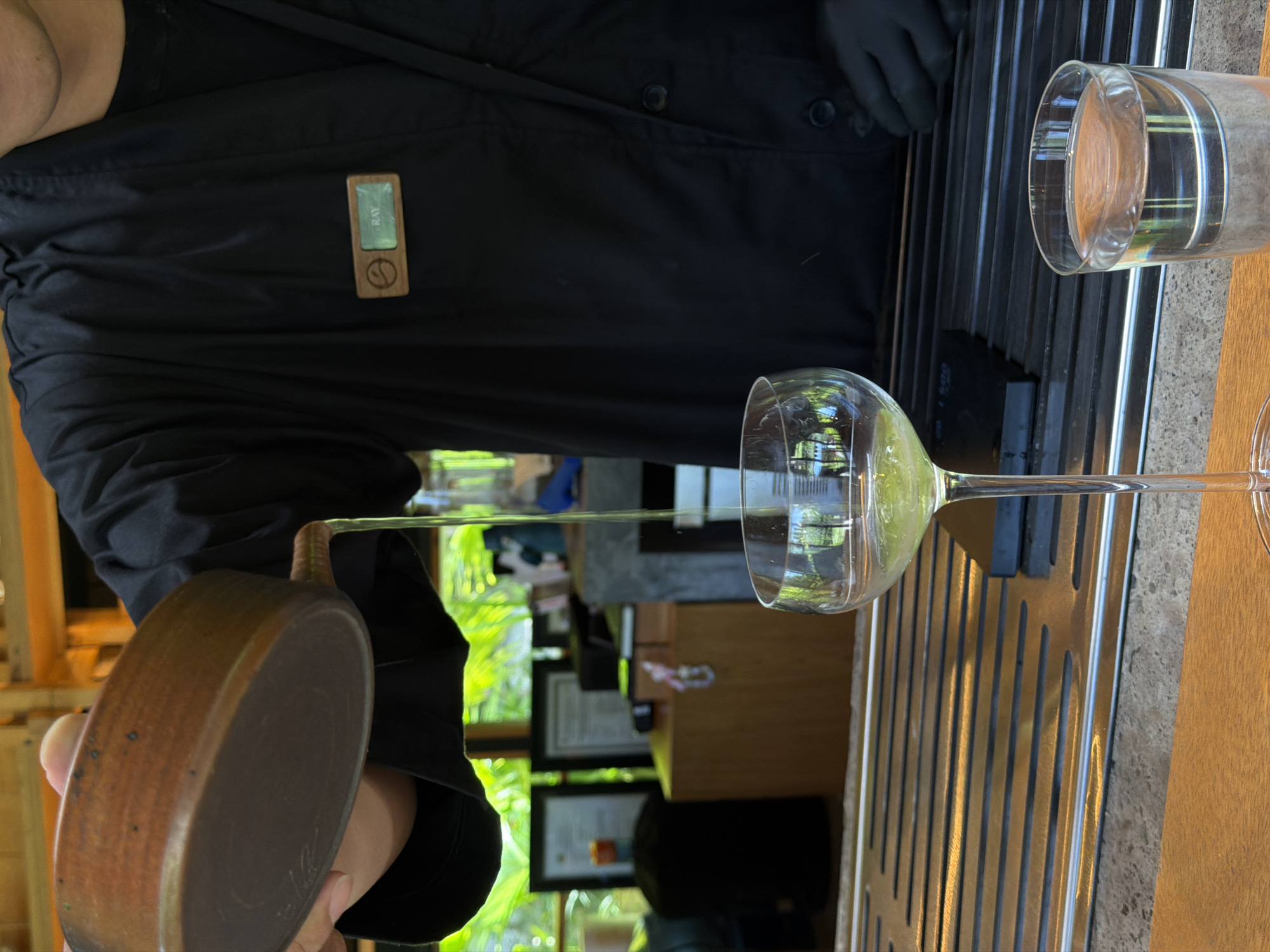 Here is a detailed description of the image:
The image captures a scene in what appears to be a restaurant or dining area in Ubud, Indonesia. A person, possibly a waiter or staff member, wearing a black shirt with a name tag that reads "BAY" is pouring a liquid from a small brown vessel into a wine glass. The liquid being poured appears to be transparent. A second glass, filled with water, is positioned on the right side of the frame, further away. The scene is set on a table with a distinct pattern of metallic bars running along it. The background reveals an outdoor view with lush greenery, partially visible through a window or opening. The overall lighting suggests it's daytime. The scene is oriented from a point-of-view perspective of someone sitting at the table.