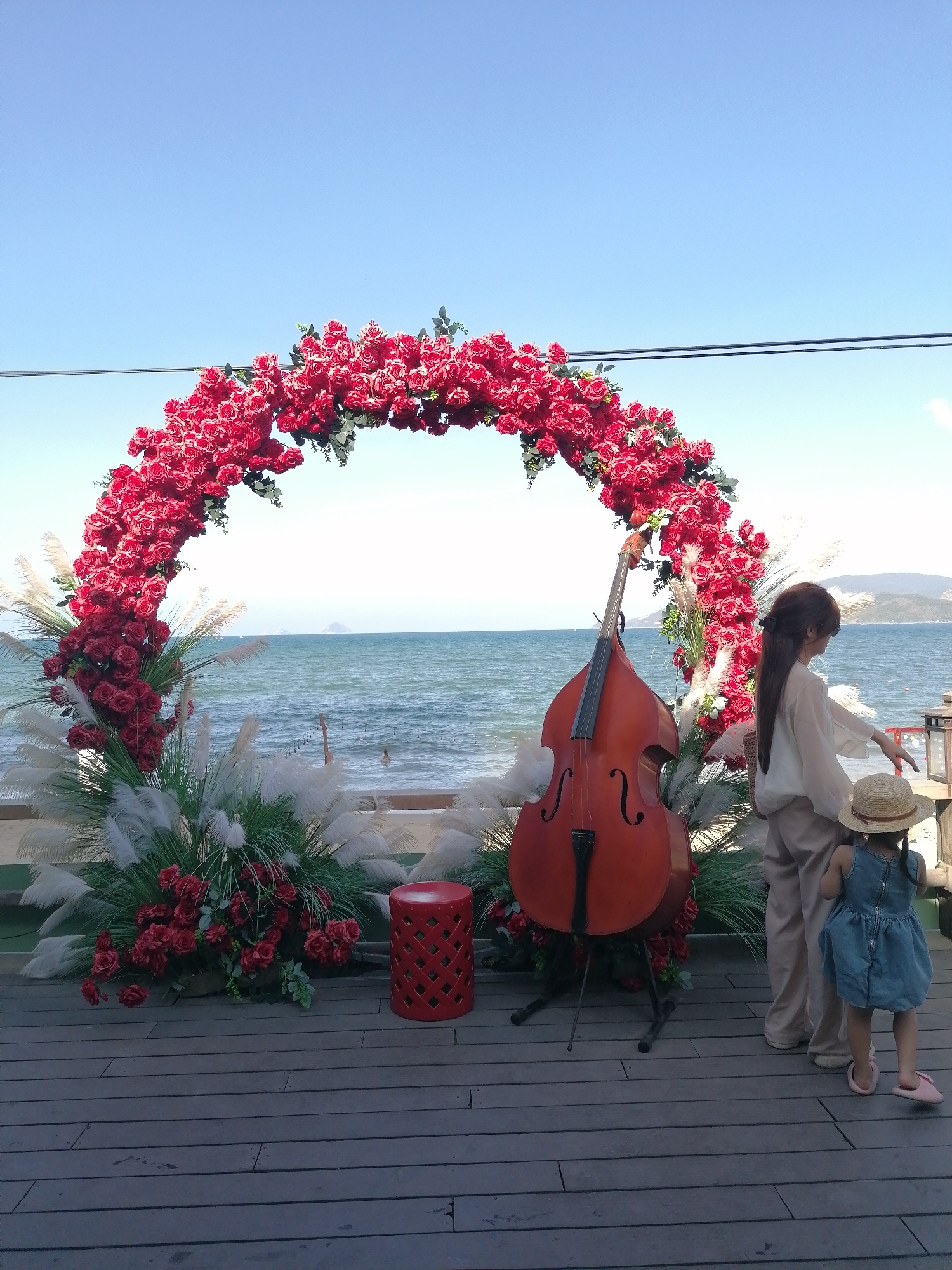 A daytime outdoor scene captures a floral archway adorned with red roses and pampas grass, set against a backdrop of the ocean and the horizon line. To the right of the archway, a woman with long dark hair, dressed in light-colored clothing, stands next to a small child wearing a straw hat and a blue dress. A large string instrument, possibly a cello or double bass, is positioned within the archway. A small red stool sits to the left of the instrument. The scene is situated on a wooden deck, possibly in Nha Trang Ward, Vietnam. The sky is blue.
