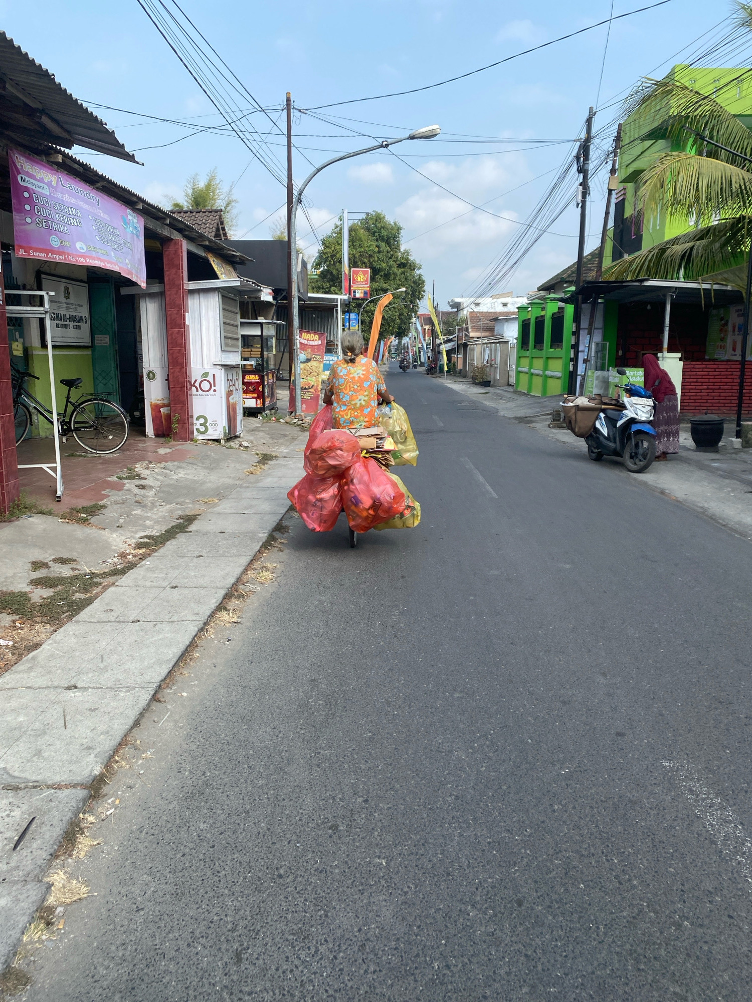 In Kediri City, Indonesia, a person on a bicycle navigates a street lined with businesses. The cyclist, from the back, is adorned in a vibrant orange and blue patterned shirt and is transporting a large load of red and yellow plastic bags, likely containing recyclables or waste, strapped to the back of the bicycle. The street is paved and appears to be a commercial area, with buildings of various colors, including green and red, lining both sides. A blue and white motorcycle is parked on the right side of the street, near a woman in a maroon hijab. Utility poles with a dense network of electrical wires are prominent features of the urban landscape. The sky is clear and blue, indicating a sunny day.