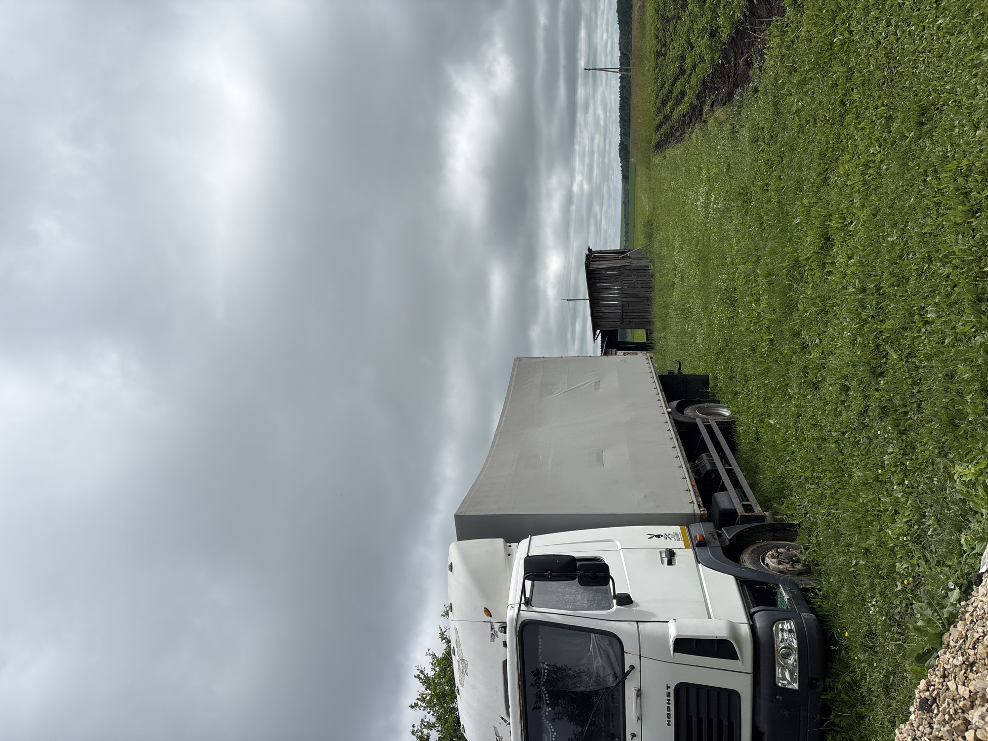 A white truck with a grey canvas-covered cargo box is parked on a grassy field in Ильпанур, Russia. To the right of the truck is a small, dilapidated wooden structure. Beyond the structure, a cultivated field is visible, with rows of plants and tilled soil. The background consists of a treeline and a cloudy sky. The image appears to be taken outdoors during the daytime.