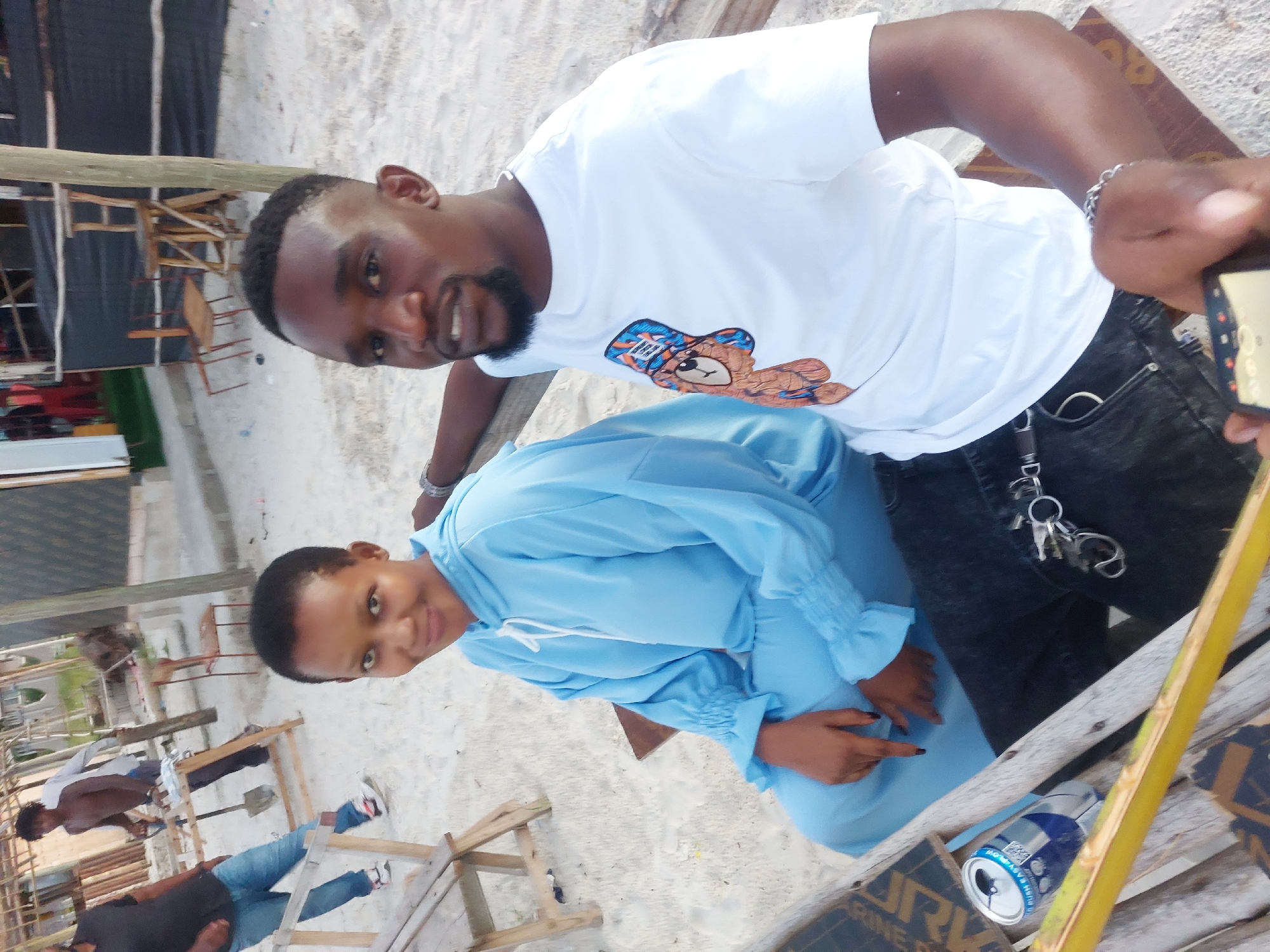 The image shows two people posing for a photo on a beach. A man with a beard is wearing a white t-shirt with a graphic of a bear. A woman is wearing a light blue dress. They appear to be sitting on or near some wooden structures, possibly part of a beachside setup. In the background, there is a sandy beach with other people and some wooden furniture. A can of soda or beer is visible near the bottom of the image. The location is likely a beach in Dar es-Salaam, Tanzania.