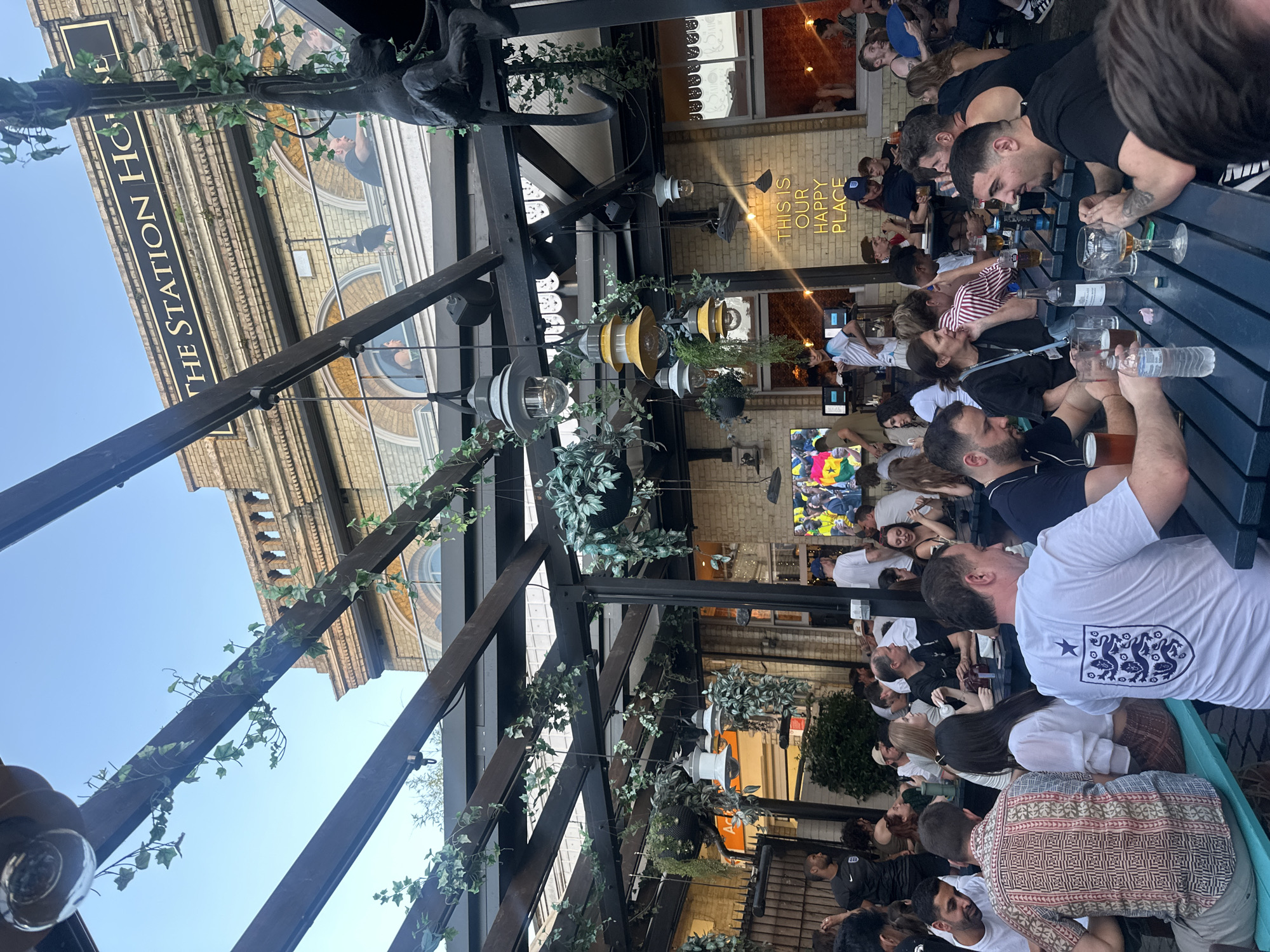 The image shows an outdoor seating area of "The Station House" pub, located in London, United Kingdom. A group of people are seated at dark blue tables, some are eating and drinking, while others are engaged in conversation. The pub has a brick facade with arched windows. The outdoor area is covered by a wooden pergola with hanging plants and lighting fixtures. A sign on the building reads "THE STATION HOUSE". Neon text on the wall reads "THIS IS OUR HAPPY PLACE". In the background, a large screen displays a football match. The people are dressed in casual attire. The overall scene depicts a lively atmosphere at a pub on what appears to be a pleasant evening.