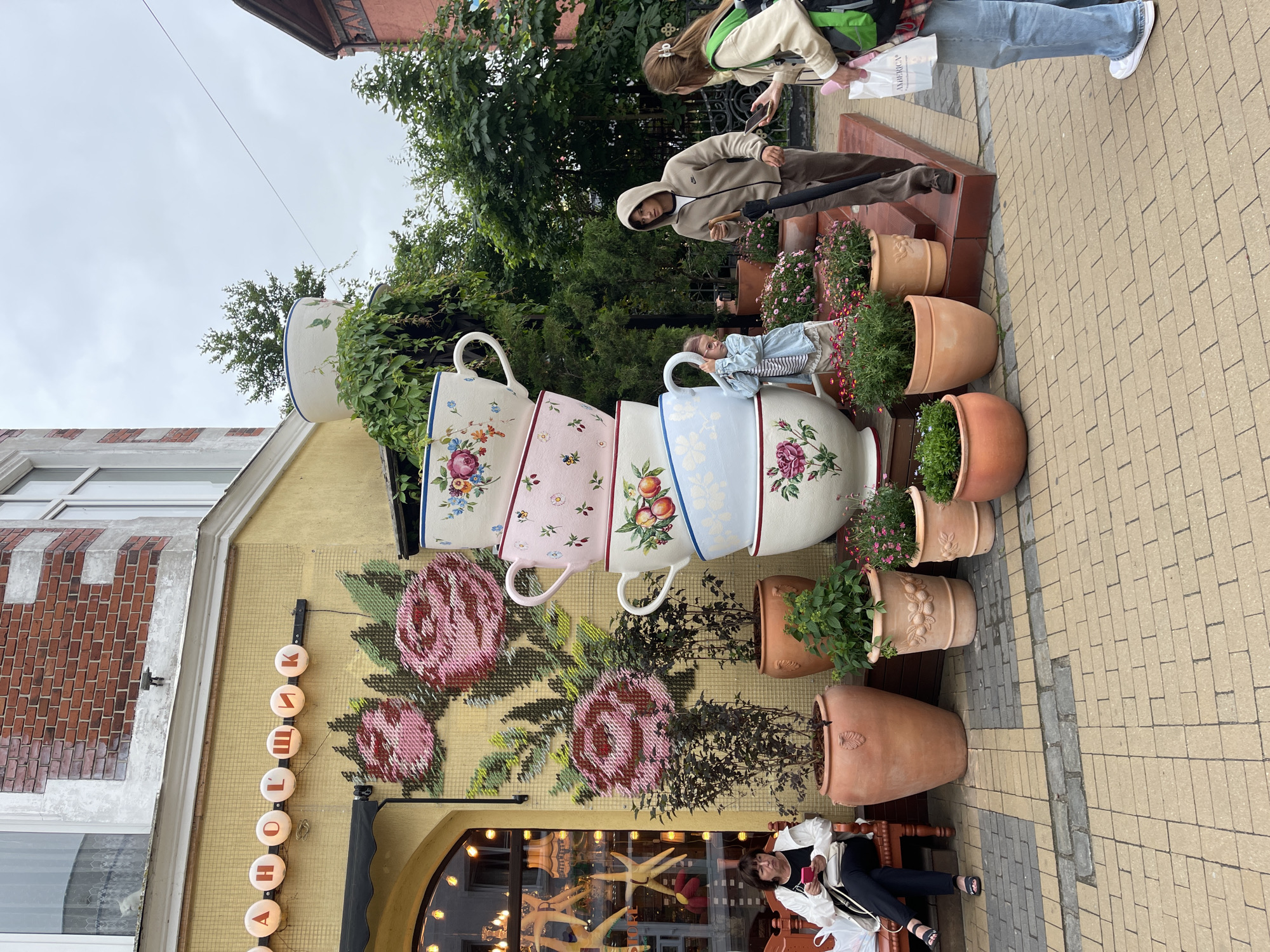 The image captures a scene in front of a yellow building with a red brick upper section, located in Zelenogradsk, Russia. A sign with circular illuminated letters, spelling "АННОЛОШНИК", is mounted above a mural of large pink roses. To the right of the building, a towering stack of oversized ceramic cups, adorned with floral and fruit motifs, dominates the foreground. A young girl in a light blue denim jacket and white skirt peeks from behind the cups. Two young adults are visible near the cups: a young man wearing a beige hooded jacket and brown pants, holding a black umbrella, and a young woman with a green backpack, wearing a light-colored jacket and blue jeans. They appear to be interacting, with the woman holding a mobile phone. On the left side of the scene, a woman in a white jacket and black pants sits on a bench, looking at her phone. Several large terracotta planters filled with greenery and flowers are arranged around the cups and the bench. The ground is paved with a mosaic of yellow and gray bricks. The sky is overcast.