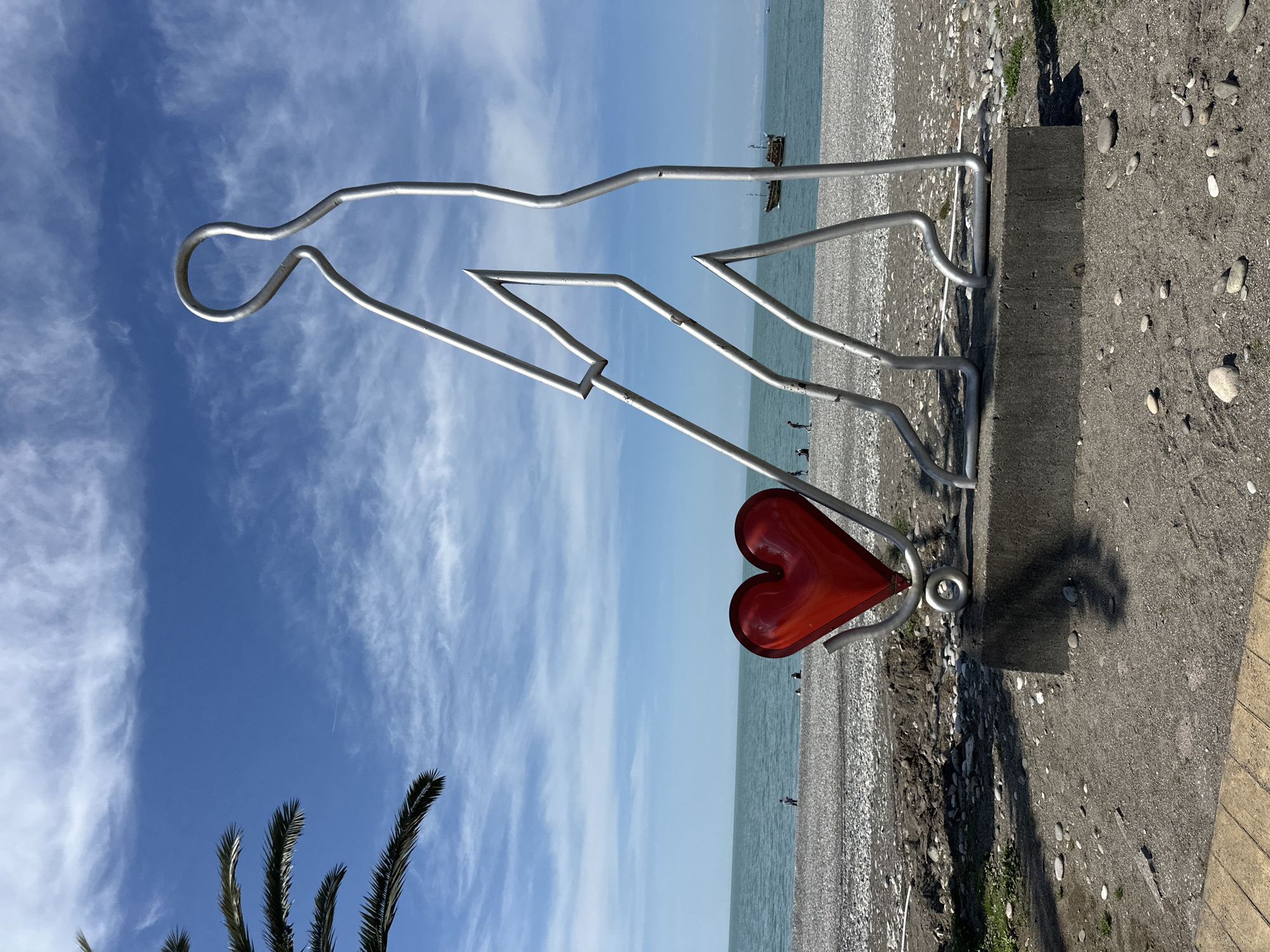The image depicts a metal sculpture of a person pulling a red heart, set against a backdrop of the Black Sea and a partly cloudy blue sky in Batumi, Georgia. The sculpture is made of silver metal rods and rests on a concrete base positioned along a pebble beach. The location is likely the Batumi Boulevard. A few individuals are visible on the beach in the distance. Some palm branches are visible in the lower left corner of the image.