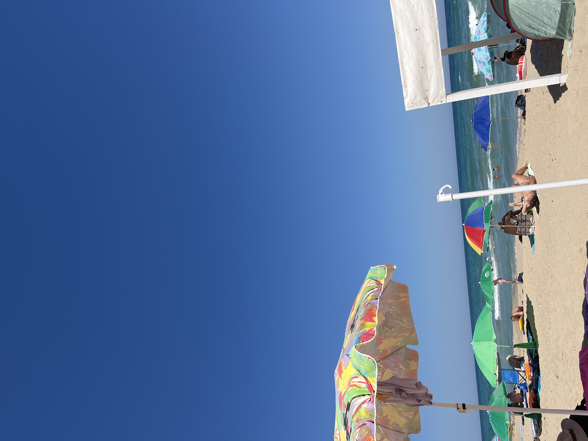 The image captures a sun-drenched beach scene in Obzor, Bulgaria, under a clear blue sky. The foreground is dominated by the vibrant, multi-colored canopy of a beach umbrella. Beyond this, the sandy beach is dotted with numerous other beach umbrellas in various colors, including green, blue, and a smaller rainbow-patterned one. Several people are visible on the beach, some relaxing on towels or loungers, while others are seated under the shade of umbrellas. A few individuals are also in the turquoise sea, enjoying the waves. To the right, a white canopy structure stands prominently, providing shade. The overall impression is a lively, typical summer day at a Bulgarian coastal resort.