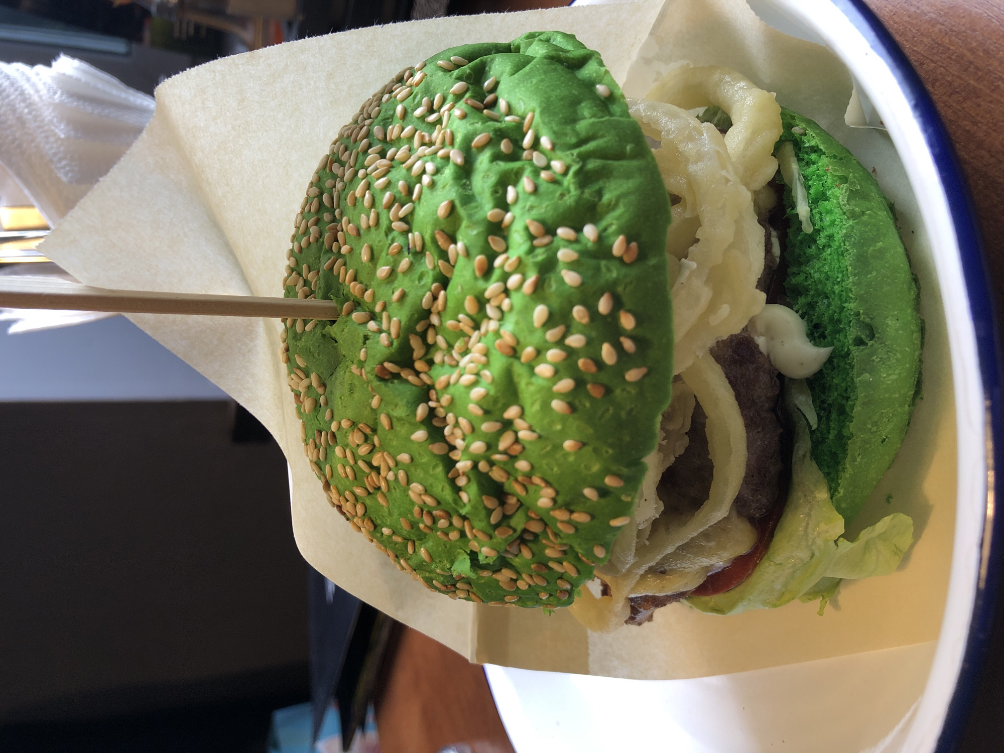 A close-up shot shows a vibrant green hamburger with sesame seeds on the bun, served in a white bowl with a blue rim. The burger is held together by a wooden skewer and is partially wrapped in a light brown paper. Visible layers include lettuce, a meat patty, onion rings, tomato sauce, and a white sauce. The setting appears to be a restaurant table with a wooden surface. The image was taken in Izhevsk, Russia.