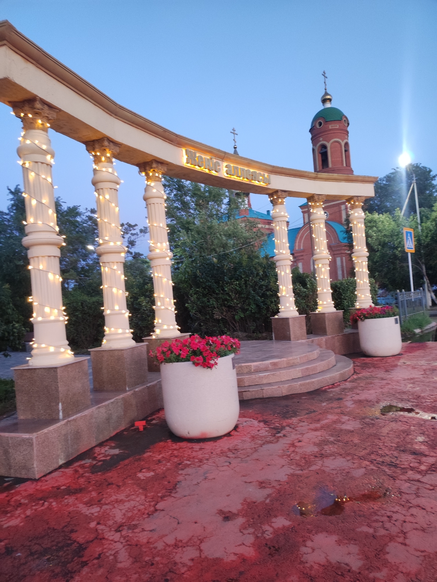 The image shows an arched structure with white columns, adorned with string lights. Behind the archway, a red brick church with a green dome and a cross is visible. In the foreground, there are two large white planters filled with red flowers. The ground is paved with a red surface that appears wet and cracked in places. A pedestrian crossing sign is visible in the background. The scene is illuminated by the string lights and a bright light source to the right, suggesting it is dusk or early evening. The location is identified as Hromtau, Kazakhstan.