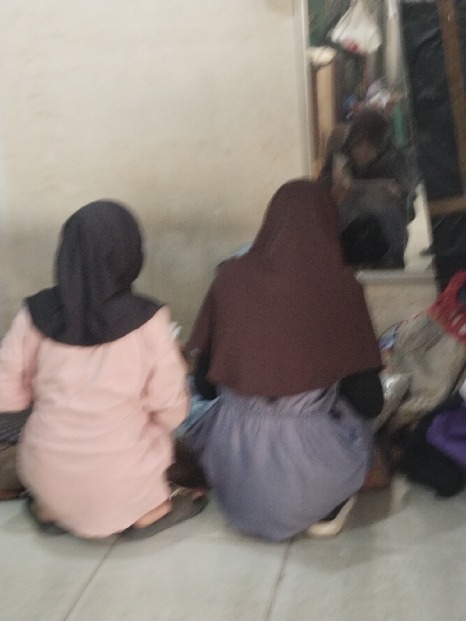 Two individuals, viewed from behind, are seated on the floor. The individual on the left wears a black hijab and a light pink garment. The individual on the right wears a dark brown hijab and a gray skirt. The environment appears to be indoors, with a light-colored wall and tiled floor visible. In the background, a reflection in what seems to be a mirror or glass surface shows more of the interior, including shelves, bags, and another person. The overall lighting is dim.