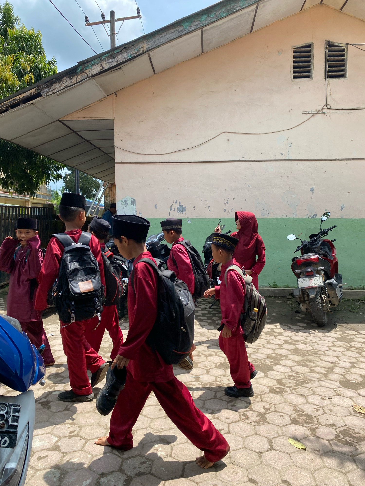 A group of male students, dressed in traditional red clothing and black caps, walk along a paved walkway outside a building. Some of the students carry black backpacks. A woman in a red headscarf and matching clothing walks behind the students. A red scooter is parked to the right of the walkway. The building appears to be aged, with peeling paint on the roofline and louvred windows. The scene is outdoors, with trees and a telephone pole visible in the background. The sky is partly cloudy. The location is identified as Karang Gading, Indonesia.