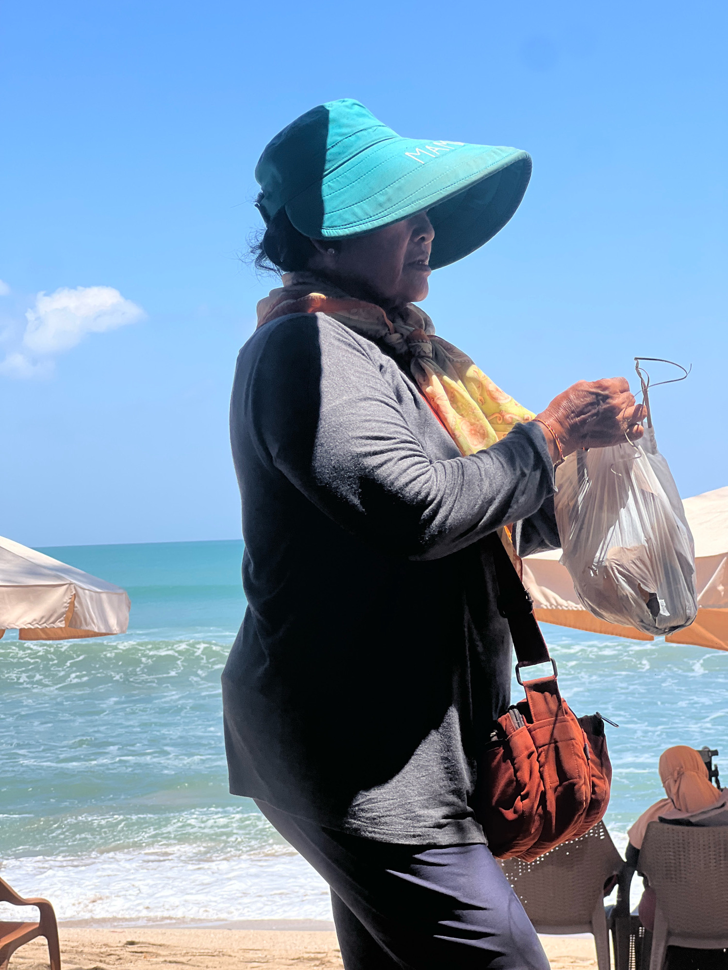 An elderly Indonesian woman, wearing a large turquoise sun hat with "MARINA" inscribed on it, a gray long-sleeved shirt, a colorful scarf, and dark pants, stands on a sandy beach in Kuta, Indonesia. She is holding a white plastic bag in her right hand and has a reddish-brown messenger bag slung over her left shoulder. The ocean with turquoise water and white waves is visible behind her, along with a pale beige beach umbrella. In the background, another person in a light-colored headscarf sits on a chair under a similar umbrella. The sky is clear and blue with a single white cloud.