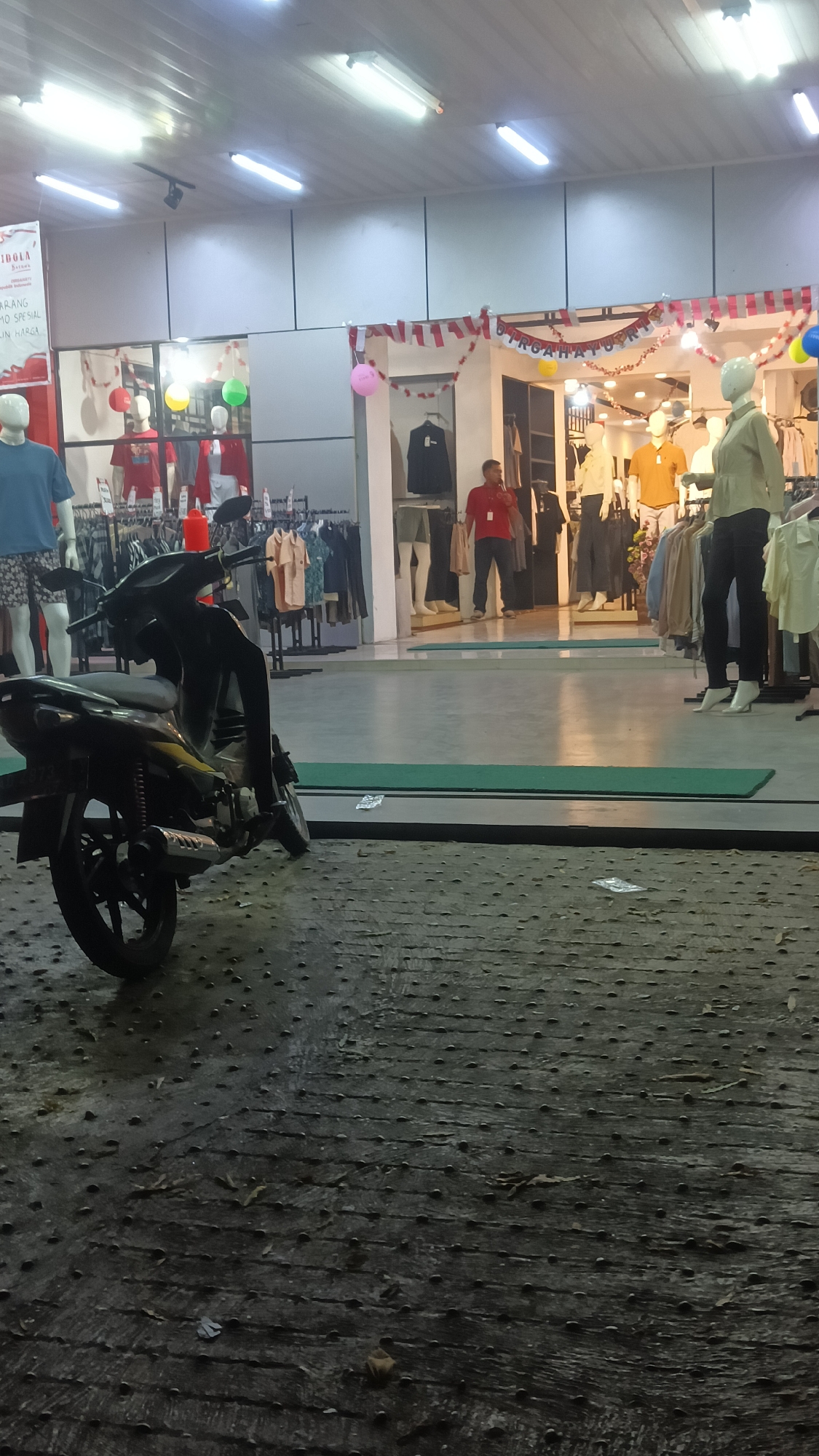The image depicts the exterior of a clothing store in Sragen, Indonesia, at night. A dark-colored motorcycle is parked in the foreground on a textured, patterned concrete surface. Beyond the motorcycle, the store entrance is visible, with mannequins displaying clothing inside. Red and white decorative garlands are hung above the entrance. Fluorescent lights illuminate the store's interior, revealing racks of clothing and a few individuals. One person, wearing a red shirt, stands near the entrance, facing away from the camera. Another individual, in a light-colored shirt, is visible further inside the store. The overall impression is of a retail establishment operating in the evening.