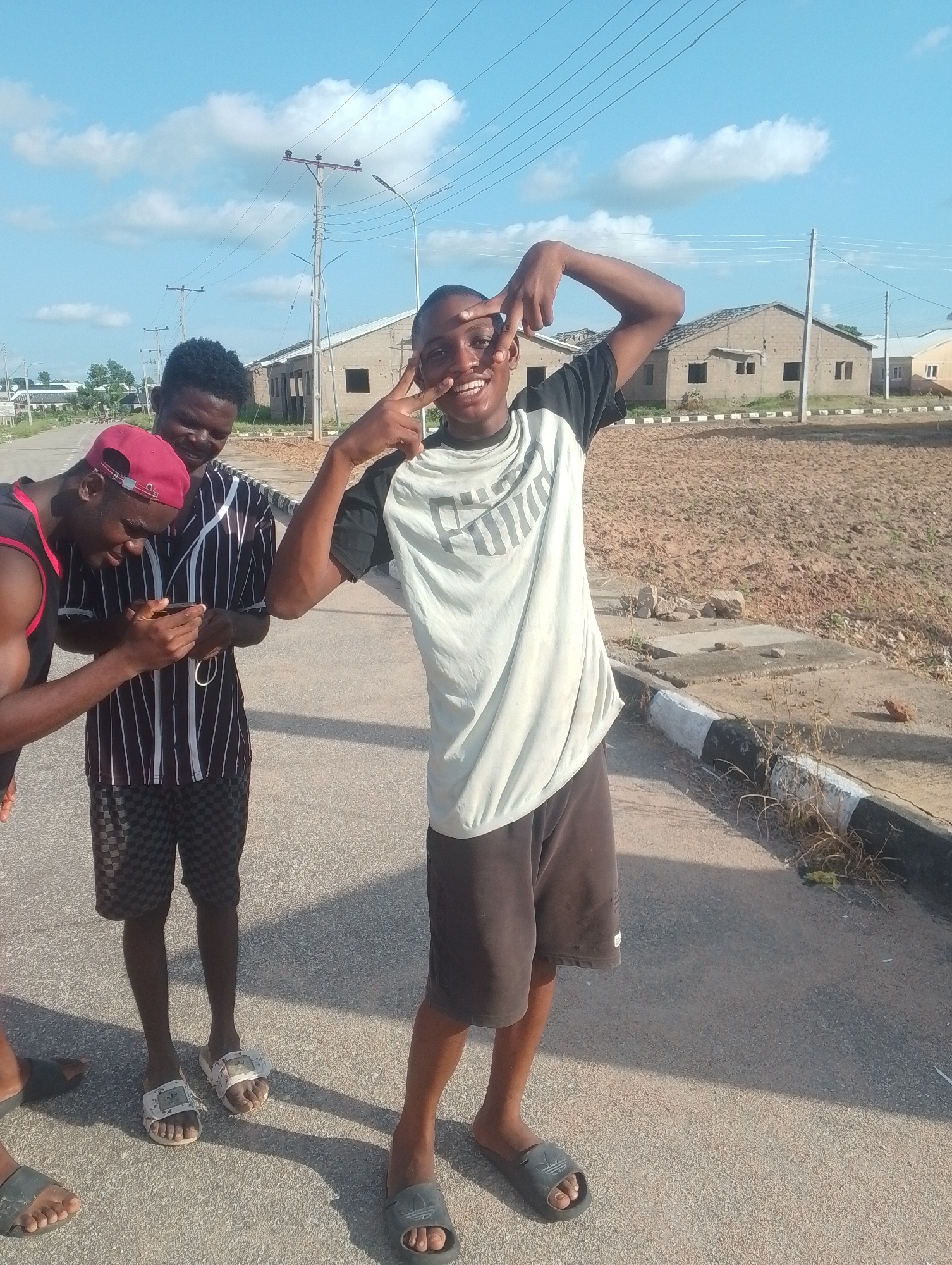 Three young men are standing on a paved road in what appears to be a developing urban area. The man in the foreground, facing the camera, is wearing a light-colored t-shirt with a dark design and dark shorts. He is making a peace sign with his right hand and smiling. To his left, another man in a striped t-shirt and checkered shorts is looking down at a mobile phone he is holding. The third man, partially visible on the far left, is wearing a red cap and a dark sleeveless shirt. In the background, there are unfinished brick buildings, power lines, and a dirt lot. The sky is blue with scattered white clouds.