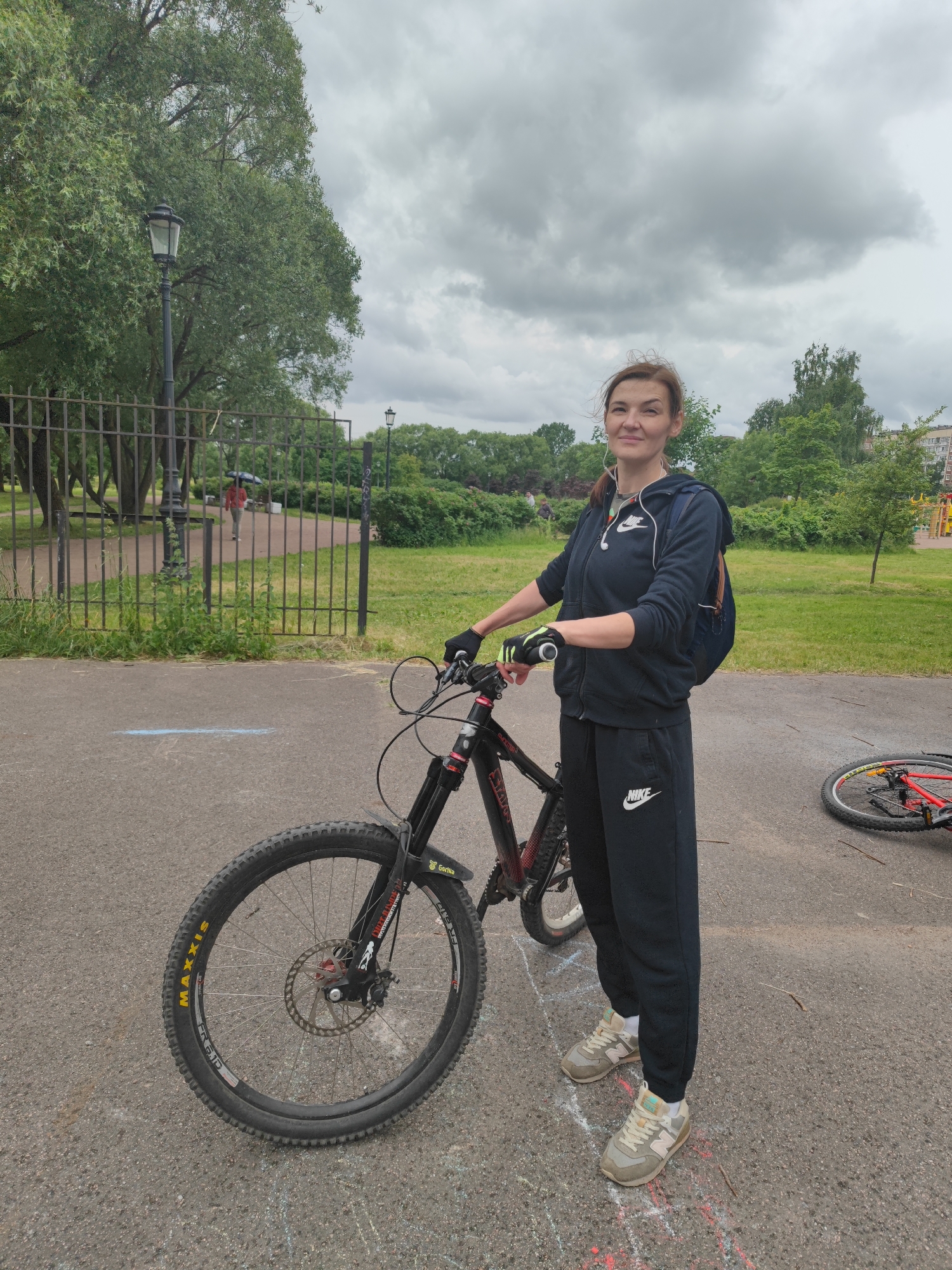 A full-shot, eye-level, outdoor photograph captures a woman standing next to a bicycle on a paved surface in a park. The woman is wearing black Nike sweatpants, a black Nike hooded jacket, black gloves, and light-colored sneakers. She has earbuds in her ears and a backpack slung over her left shoulder. Her right hand is on the handlebars of the bicycle, which is black with red accents and has a "Maxxis" tire. Another bicycle, red and white, is lying on its side in the background to the right. The background features a metal fence, trees, and a cloudy sky. A lamppost is visible near the fence, and a person with an umbrella is walking in the distance beyond the fence. The ground has chalk markings, including squares and lines. The overall setting suggests an urban park environment.