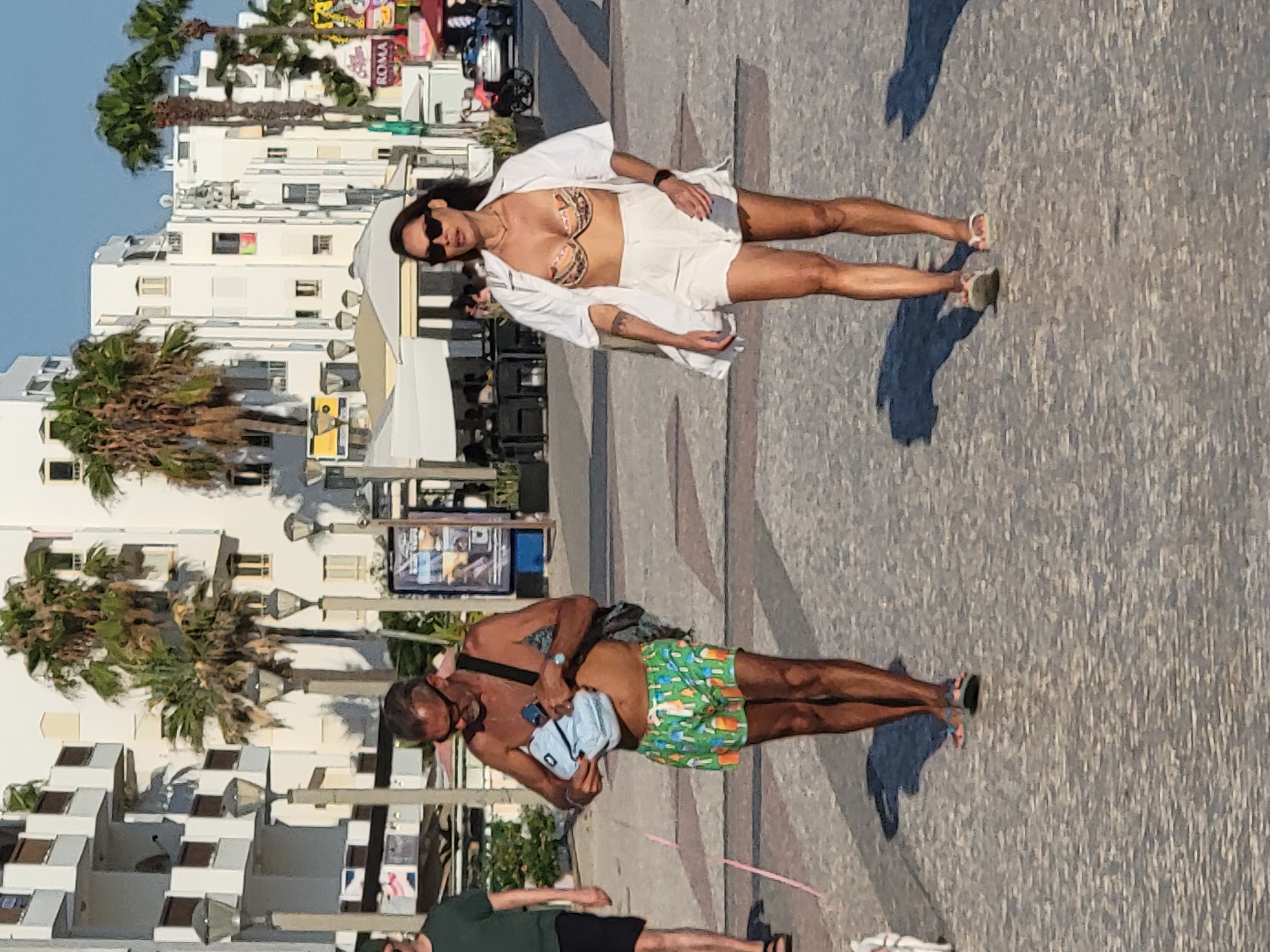 In Quarteira, Portugal, two people are seen walking on a sunny day. A woman in a white unbuttoned shirt and a patterned bikini top, paired with white shorts, walks ahead. She wears sunglasses and flip-flops. Behind her, a man in green patterned swim shorts and a dark strap across his chest is visible. He carries a light blue pouch and wears a watch on his left wrist. Another person, wearing a dark green t-shirt and black shorts, is partially visible in the foreground. Palm trees and white buildings line the backdrop, suggesting a coastal resort area. The scene appears to be a leisurely stroll on a paved promenade under clear skies.