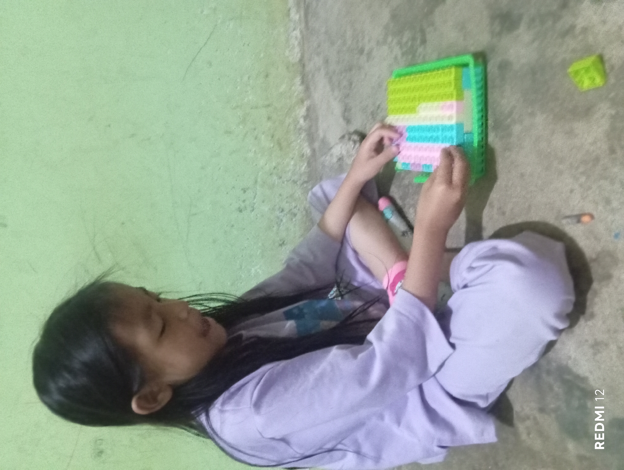 A young girl with long, dark hair is sitting on a concrete floor, playing with a colorful building block toy. She is wearing a light purple t-shirt and matching pants. The background consists of a light green wall, possibly painted concrete, with some faint marks and scratches. The lighting appears to be indoor, possibly artificial. The scene is likely a residential setting within a home in Pagar Alam, Indonesia. The girl is focused on manipulating the blocks, which are arranged in a tray. A small, lime green building block rests on the floor to the right of the tray. The overall atmosphere is one of quiet activity.
