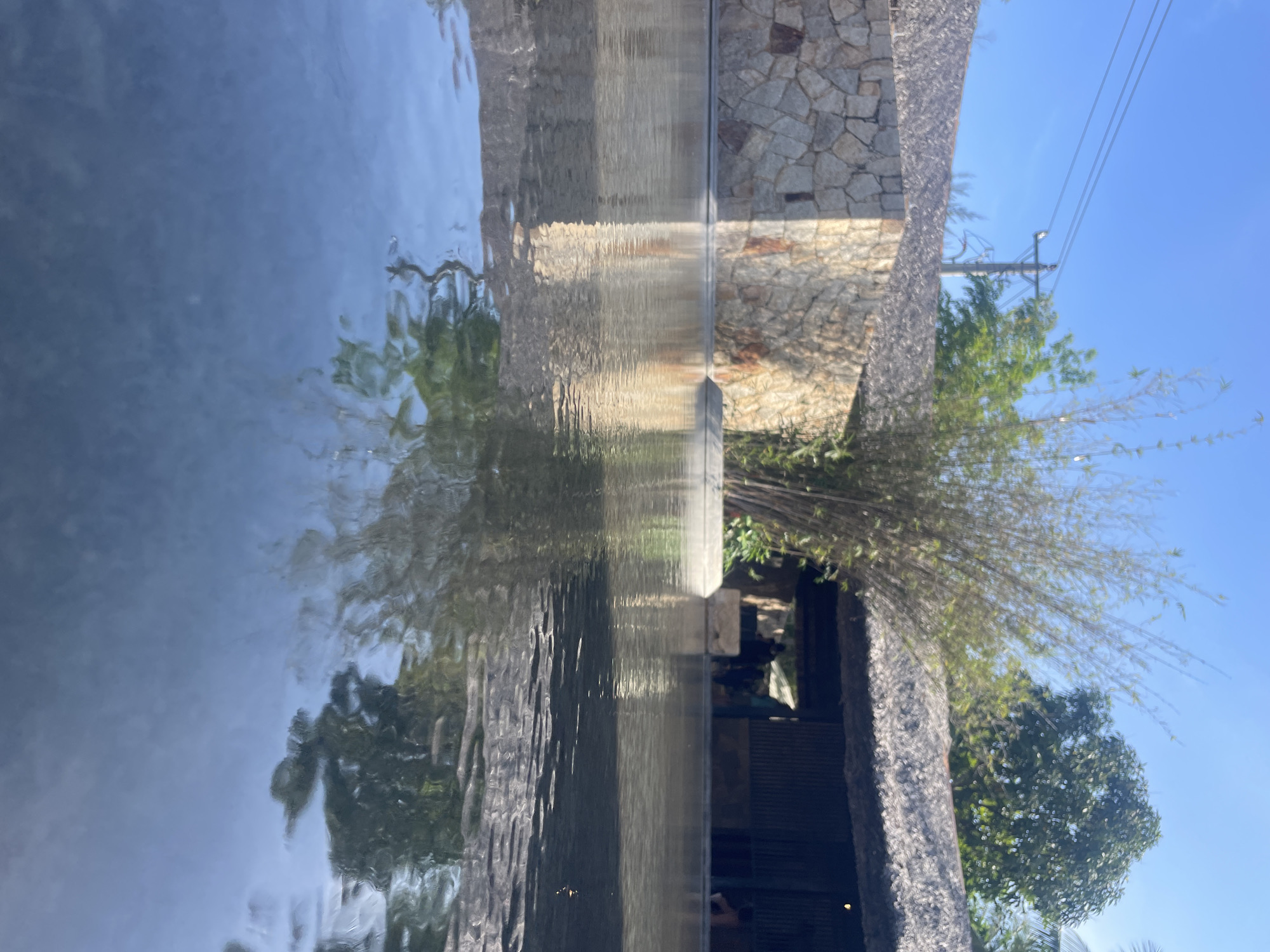 The image shows a structure partially submerged in water, possibly a building or architectural feature with a stone wall and a thatched roof. The water's surface reflects the surroundings, creating a distorted image of the sky, trees, and the structure itself. The presence of power lines and foliage suggests an outdoor setting, potentially near a coastal area. The image could be taken in Nha Trang, Vietnam due to the architecture.