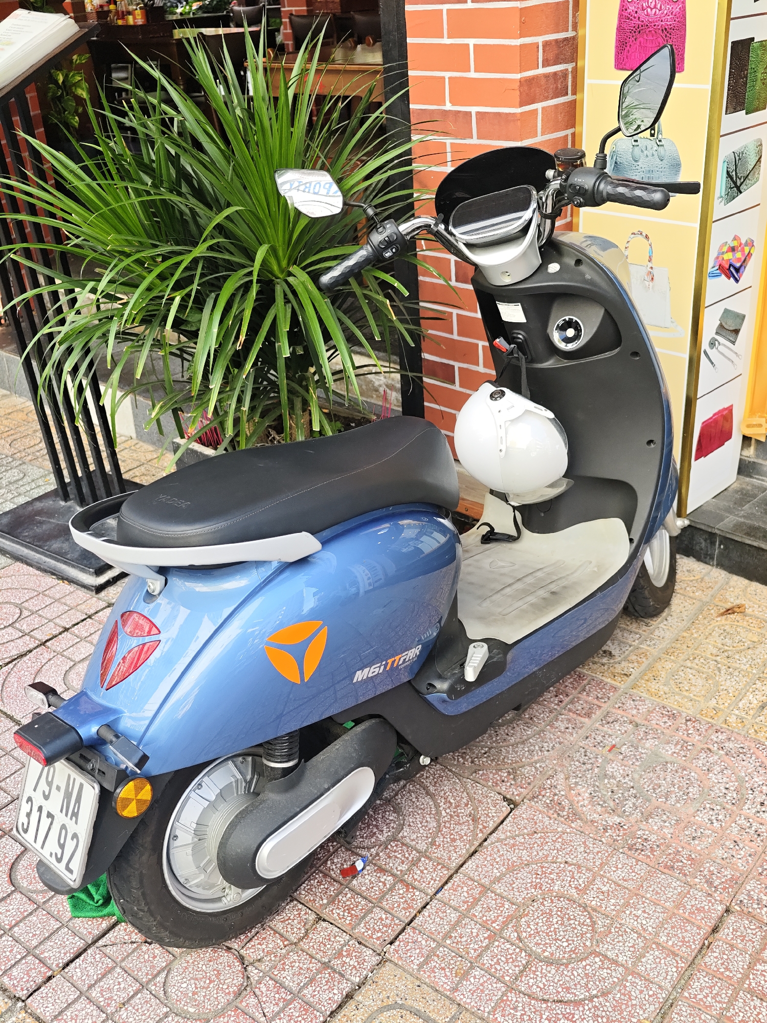 A blue electric scooter is parked on a tiled sidewalk in front of a building with a brick facade. The scooter has a black seat, a white helmet hanging from the handlebars, and a license plate that reads "79-NA 317.92". There is a large green plant to the left of the scooter, and a display of handbags and accessories is visible in the background. The scooter appears to be in a commercial area, possibly a shop or market, in Nha Trang Ward, Vietnam.