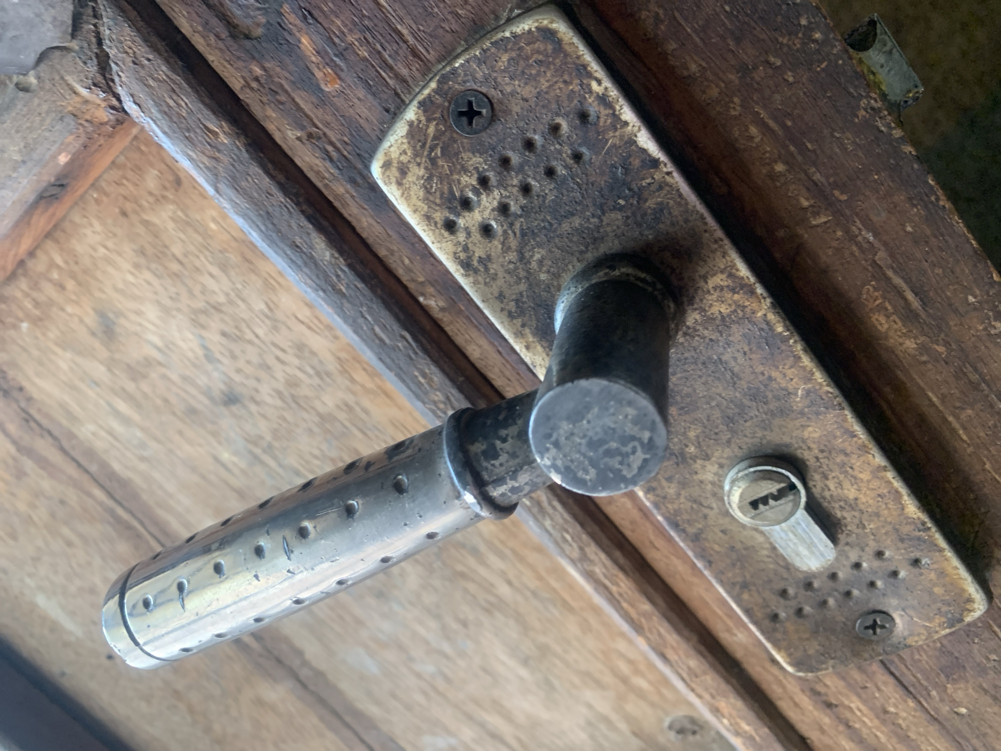 A close-up, high-angle shot shows a weathered, brass-colored door handle and lock plate mounted on a rustic wooden door. The door handle is metallic, cylindrical, and perforated with numerous small holes. The lock plate has a keyhole with a jagged-shaped opening and is secured by two screws. The wood of the door appears old and worn, with visible grain and some damage around the edges. The scene is indoors or sheltered, as indicated by the lighting and the focus on the door mechanism. There are no people or animals visible in the image. The image appears to be a photograph taken in the city of Jalingo, Nigeria.