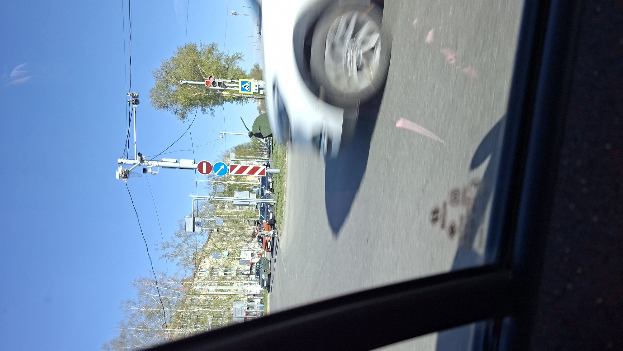 The image, taken from inside a vehicle, shows a street scene in Chaikovsky, Russia, identified by distinctive road signs. A white car is partially visible, appearing in motion and positioned in the right-hand lane. A double traffic light structure is visible in the background, with a railroad crossing warning sign and a pedestrian crossing sign. Other vehicles are further down the road. The sky is blue, and buildings are visible in the background.