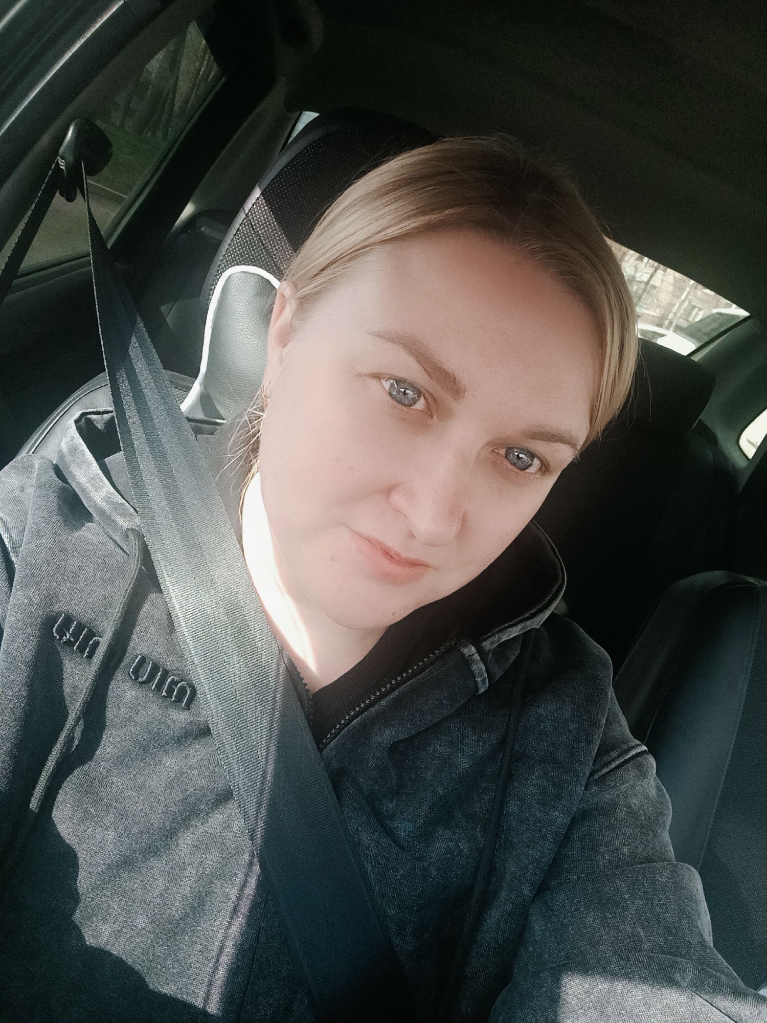 A blonde woman with blue eyes, appearing to be in her 30s or 40s, is captured in a selfie taken inside a car. She is seated, likely in the driver's or front passenger's seat, with a black seatbelt diagonally across her chest. She wears a dark grey or charcoal zip-up hoodie with a slightly distressed or worn texture. Sunlight illuminates the left side of her face and shoulder, indicating a bright daytime setting.
The car's interior is visible, including a dark seat, a headrest with a textured pattern, and part of the car door and window frame. Through the car window, a blurred outdoor scene with green foliage and what appear to be trees suggests an external environment, possibly rural or suburban.
The woman's expression is neutral, with her gaze directed towards the camera. On the front of her hoodie, embroidered text is visible, which appears to spell "UIM" on the left side of her chest, and a mirrored "UIM" on the right side. The overall scene conveys a casual moment during travel or a stop.