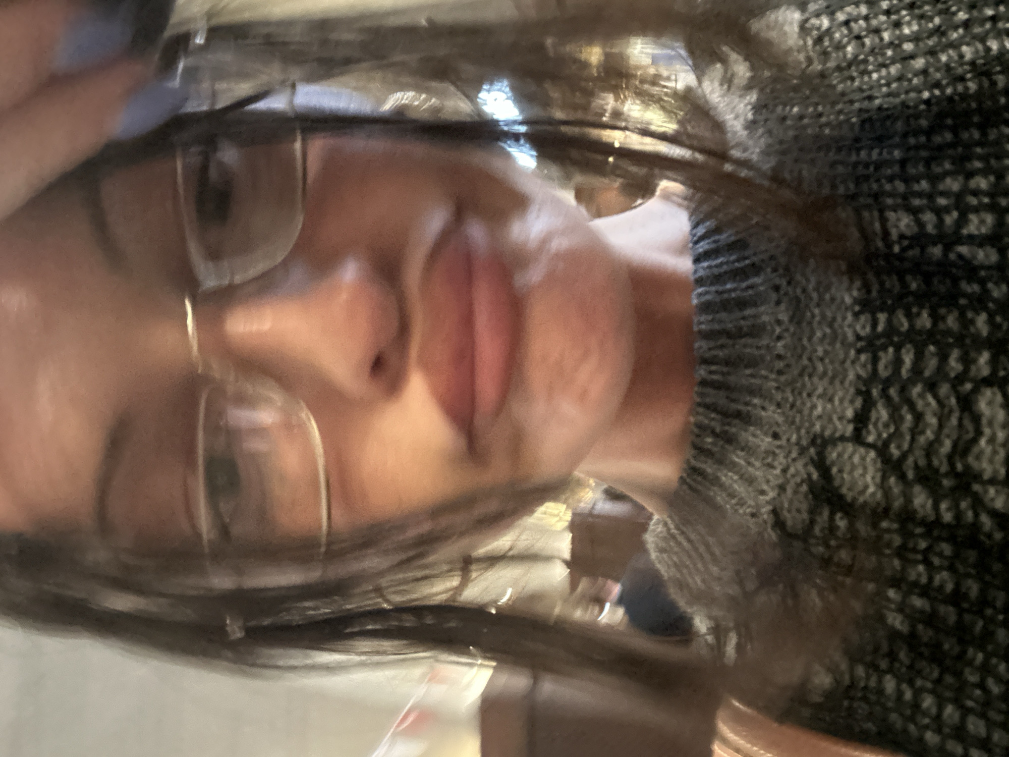A close-up, slightly blurred selfie of a person with glasses. The person is wearing a dark grey, textured knit sweater. The background is indistinct but appears to be an interior space. There is no clear indication of the location being London, United Kingdom.