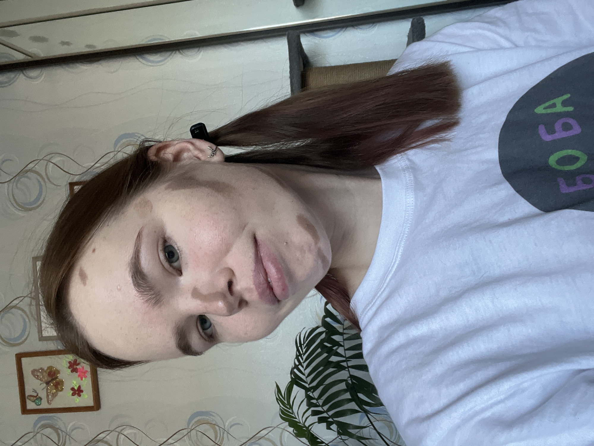 A fair-skinned woman is indoors, wearing a white T-shirt with a dark grey circle containing the word "BOBA" in green and purple letters. Her brown hair is pulled back in a ponytail held by a black clip. The background includes wallpaper with swirling patterns, a framed picture with a butterfly and flowers, and a potted plant with green leaves. The woman appears to have birthmarks or skin pigmentation variations on her face. The image was taken inside an interior, and is from the city of Chaikovsky, Russia.