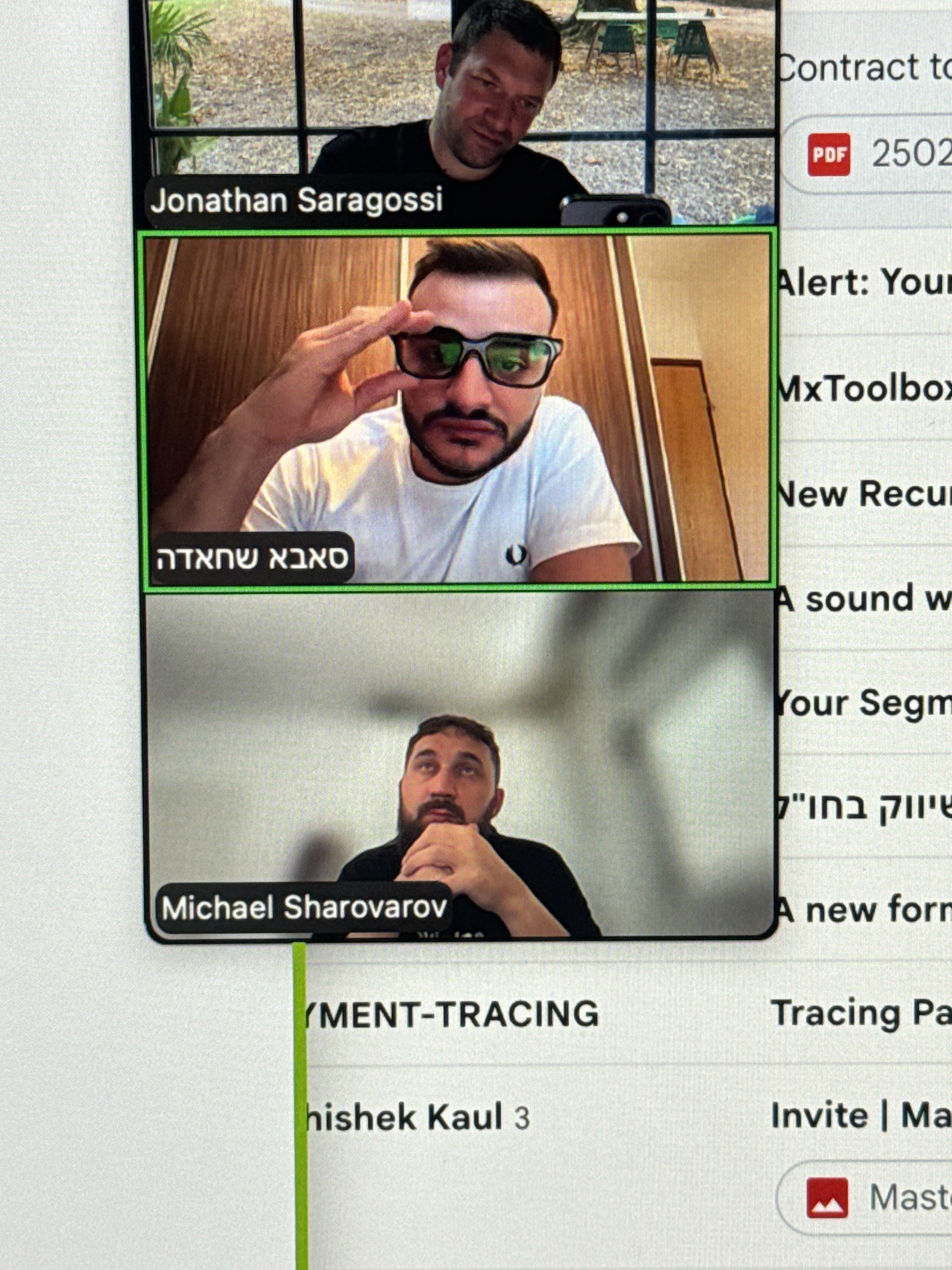 The image is a screenshot of a video conference call, likely from a platform like Zoom or Google Meet. It shows three participants in separate video panes. The top pane shows a man in a black t-shirt looking down and to the side, with an outdoor background visible behind him. The middle pane shows a man wearing glasses and a white t-shirt, with his hand raised to his forehead. Below his video pane, a black label reads "סאבא שחאדה" in Hebrew. The bottom pane shows a man with a beard wearing a black t-shirt, looking upwards with his hands clasped. Below his video pane, a black label reads "Michael Sharovarov". To the right of the video panes, text is visible, including "Contract to PDF 2502", "Alert: Your", "MxToolbox", "New Recur", "A sound w", "Your Segm", "שיזזוק בוח" in Hebrew, "A new form", "YMENT-TRACING", "Tracing Pa", "hishek Kaul 3", and "Invite | Ma" with a red Mastercard logo. The image does not contain any information or visual cues related to the city of Erba, Italy.