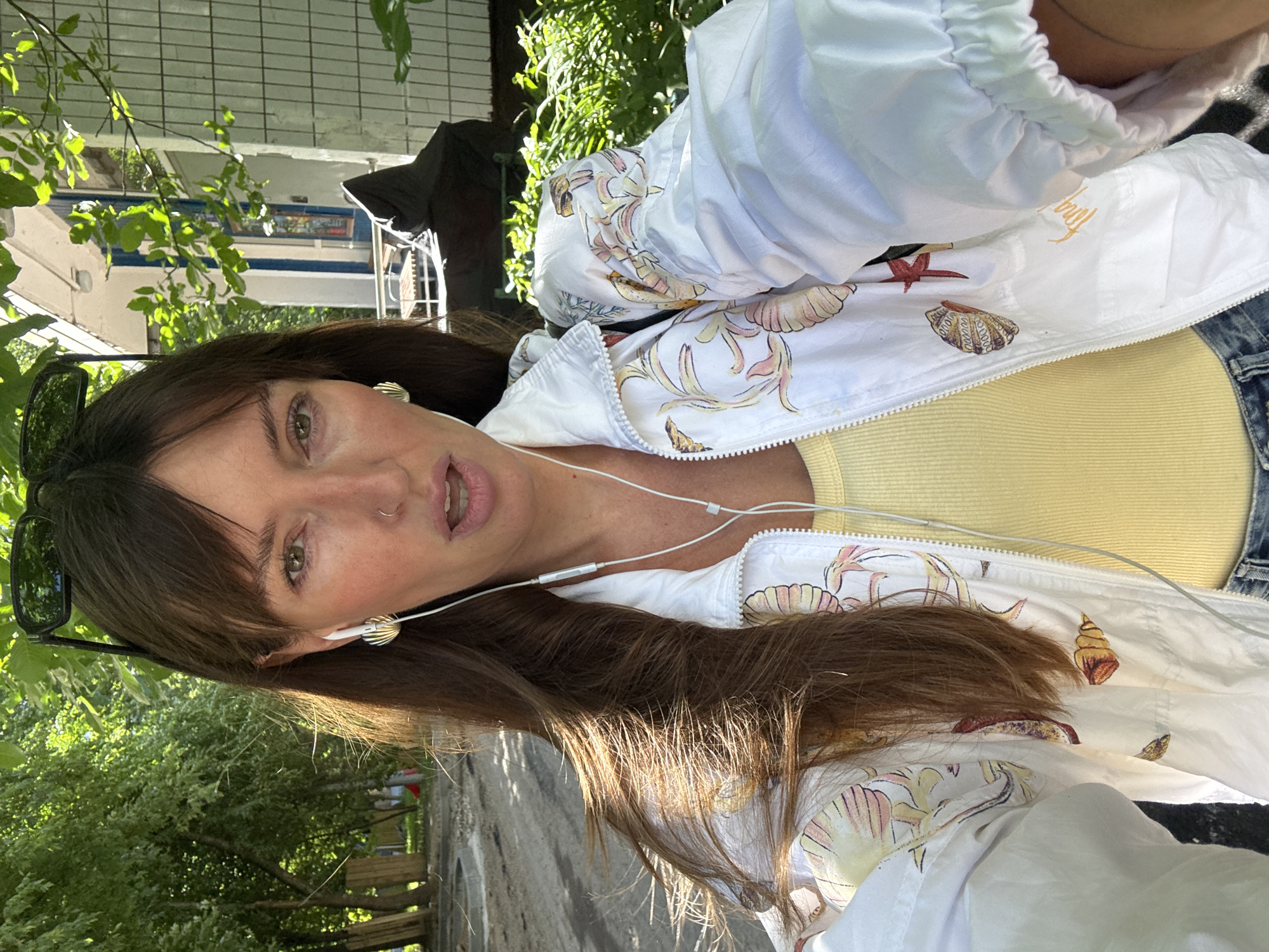 A woman with brown hair and bangs is taking a selfie outdoors. She is wearing a white jacket with a seashell print, a yellow tank top, and sunglasses on top of her head. She has white earbuds in and a nose ring. The background shows trees and a building with blue and white accents. The scene appears to be in a residential area, possibly Moscow, Russia.