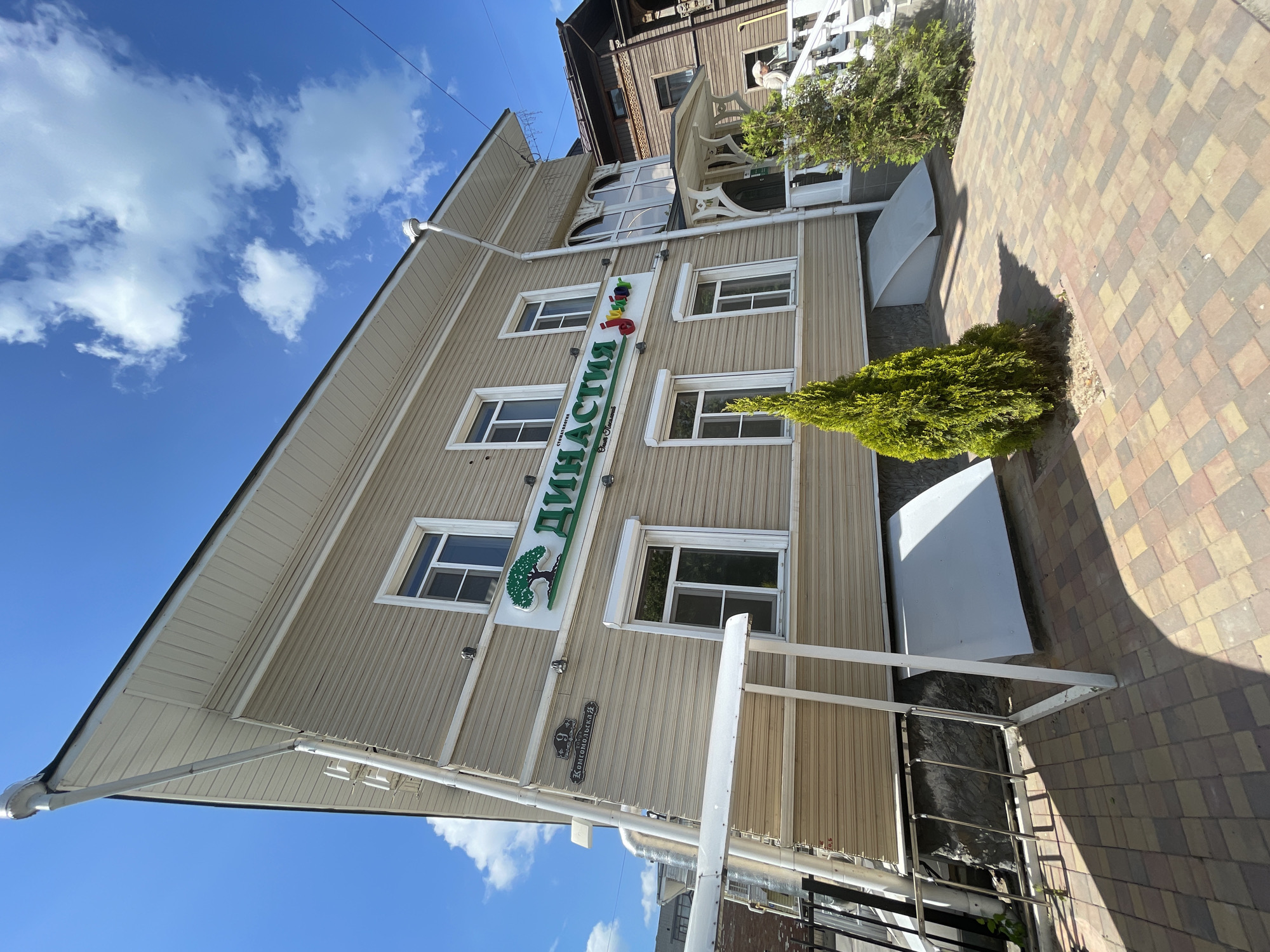 The image shows a two-story building with light-colored horizontal siding. A large green sign with a tree graphic and the Russian word "Династия" (Dynasty) is mounted above the first-floor windows. Below "Династия", smaller text reads "Стоматология" (Dentistry) and "Опыт. Успех." (Experience. Success.). To the right of "Династия" is a smaller, colorful sign that says "Junior". The building has several windows on both floors, some with white trim and roller shutters. A street sign is visible on the left side of the building, reading "9 Консомольская" (9 Komsomolskaya). A small, conical evergreen tree stands in front of the building, casting a shadow on the patterned brick pavement. In the background, a portion of a darker, wooden building is visible, and a person in a light-colored jacket is descending stairs on the right side of the image. The sky is blue with scattered white clouds. This building is located at 9 Komsomolskaya Street in Vologda, Russia, which houses a dental clinic named "Dynasty".