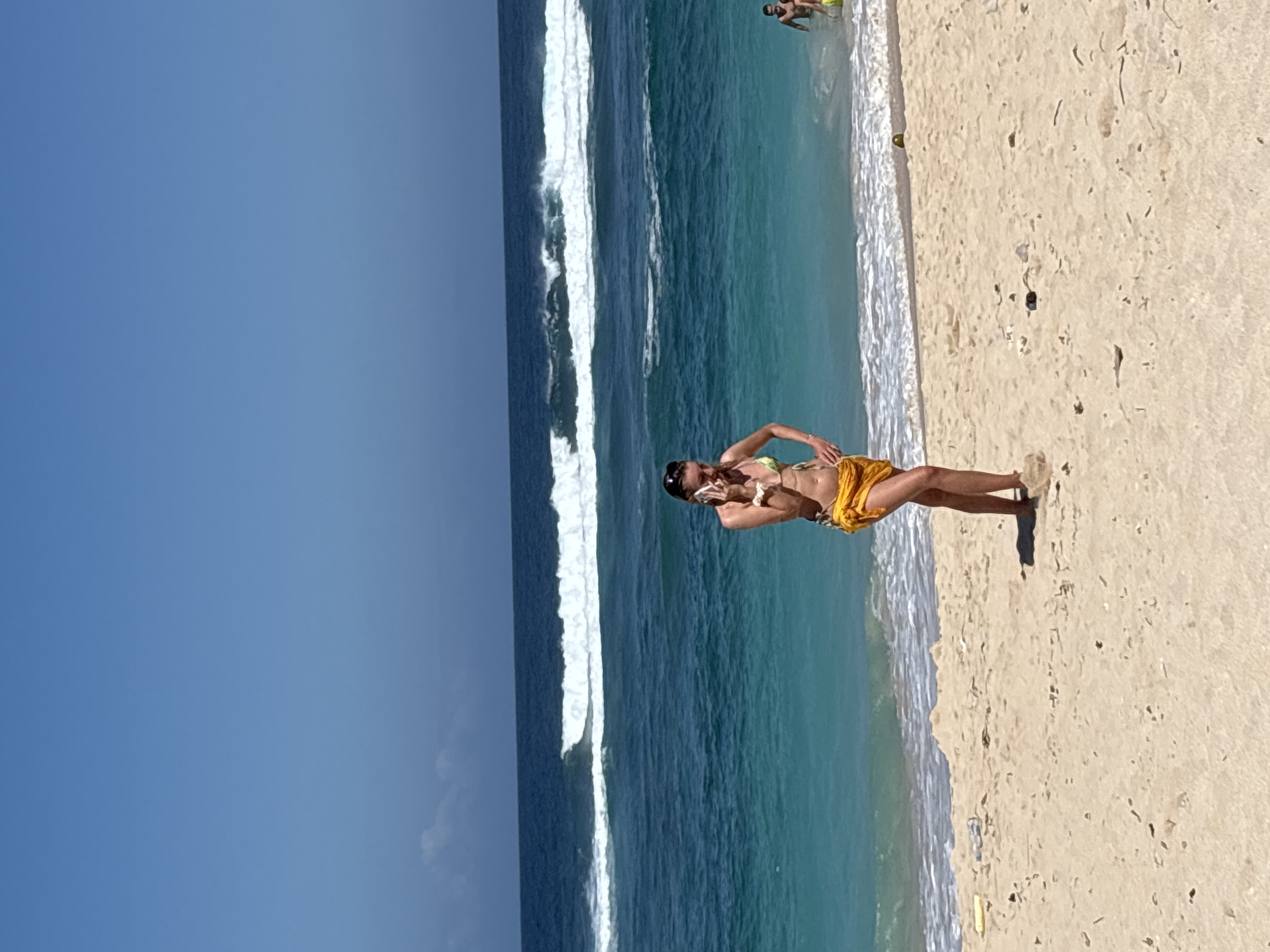 A woman in a yellow bikini and a yellow sarong stands on a sandy beach in Kuta Selatan, Indonesia, holding a smartphone to her ear. She is facing the ocean, which has large waves breaking. The water is a vibrant turquoise near the shore and a deep blue further out. The sky is clear and blue. In the background, other beachgoers are visible in the water and on the sand. The sand is light beige and appears to have some debris scattered on it. The overall scene depicts a sunny day at the beach with moderate waves.