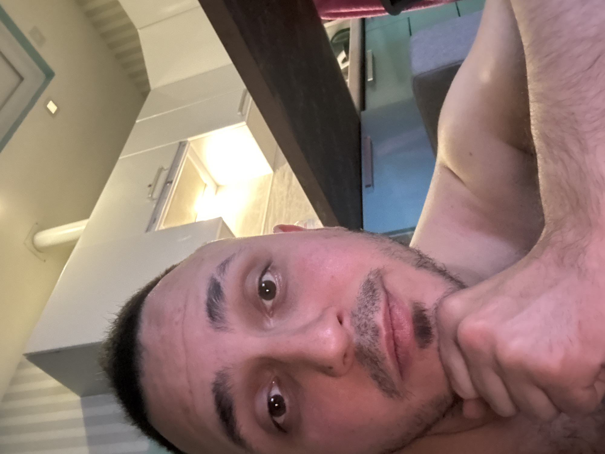 A man is depicted in a selfie-style photograph, seemingly indoors. His face is prominent, with dark hair, brown eyes, and stubble. His hand is positioned near his face. The background includes kitchen cabinets, potentially located above the subject, and a partially visible dark beam or support structure. The image lacks identifying details to associate it with Saint Petersburg, Russia. No other people, subjects, or actions are evident in the photo.