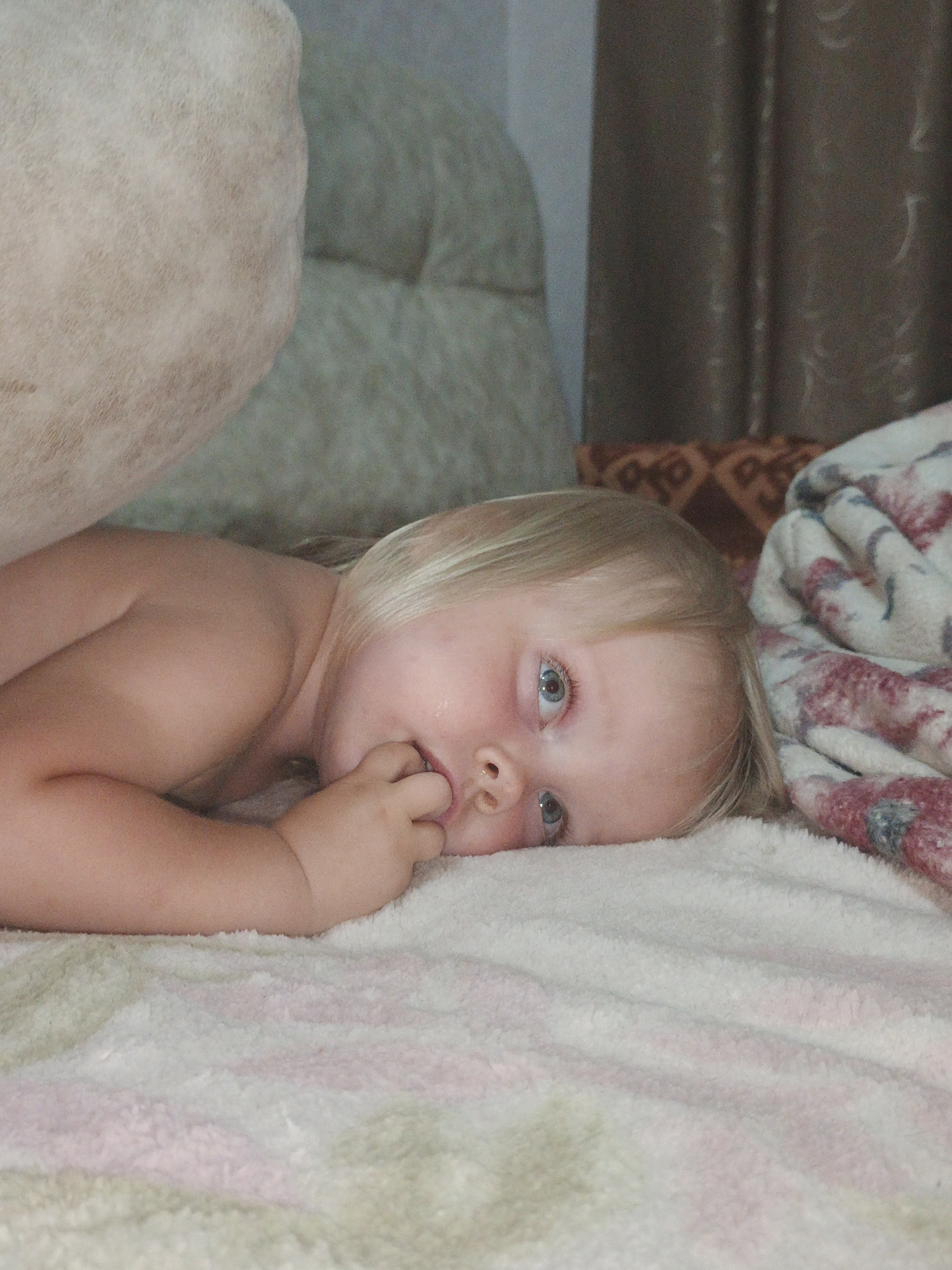 A young child with blonde hair is lying on a white, fuzzy blanket. The child is on their side, facing the camera, with their right hand in their mouth. Their eyes are open and looking towards the camera. The child's body is partially obscured by a large, light-colored object on the left, which appears to be a cushion or piece of furniture. In the background, there are brown curtains and a patterned fabric. The lighting is dim, suggesting an indoor setting. The location is not specified.