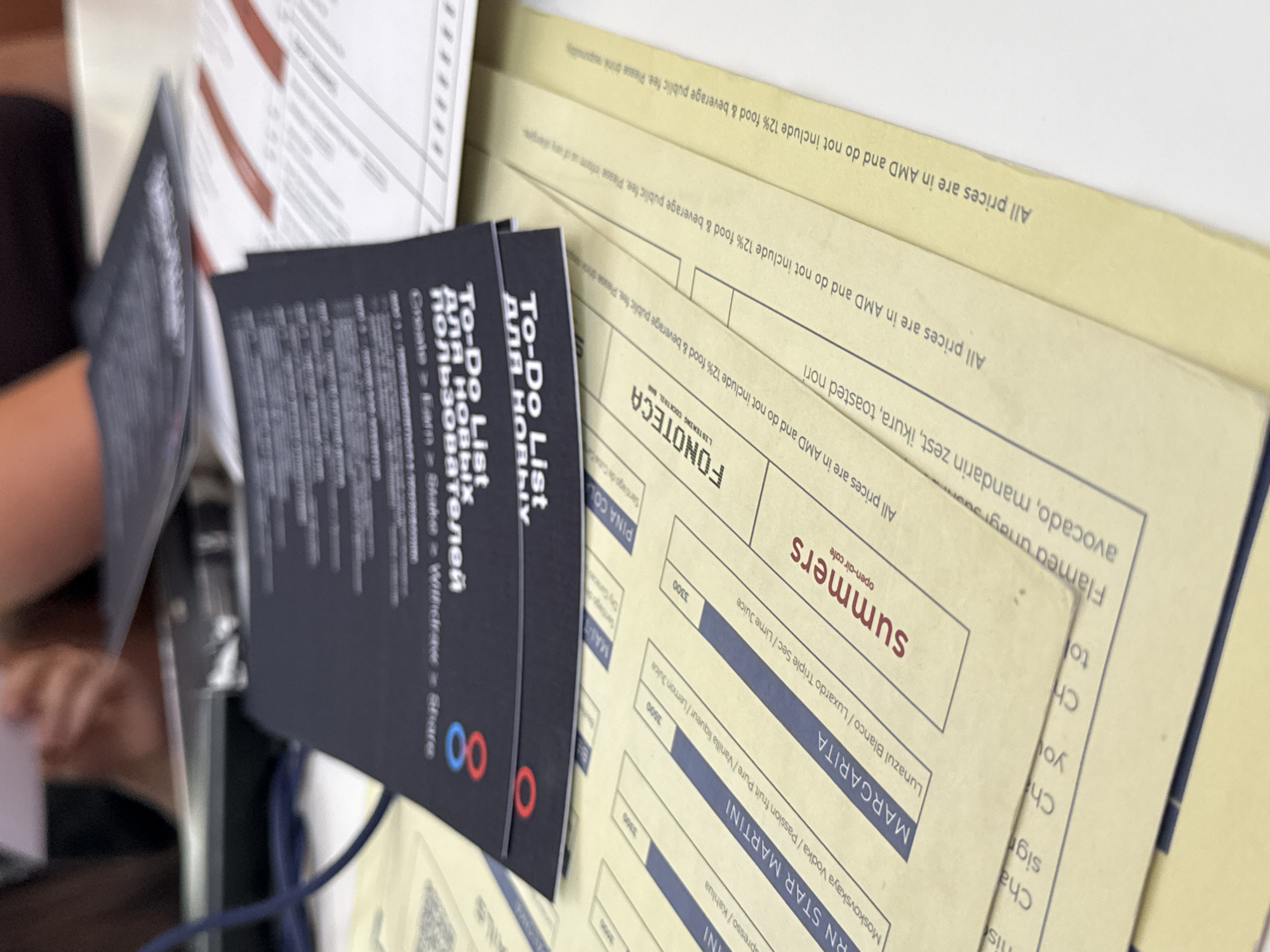 The image shows a close-up of several papers or menus stacked on top of each other, likely at a cafe or restaurant. The visible documents include a "To-Do List for New Users," translated into Russian, and a menu from "FONOTECA Listening Cocktail" in Yerevan, Armenia. The menu features cocktails like "RN STAR MARTINI," "MARGARITA," and "summers." Prices are listed in AMD (Armenian Dram) and exclude a 12% food and beverage public fee. One menu mentions "Flamed unagi, avocado, mandarin zest, ikura, toasted nori" as an ingredient. The environment seems to be a tabletop setting in a casual dining establishment. Part of a person's arm and hand is seen in the background.