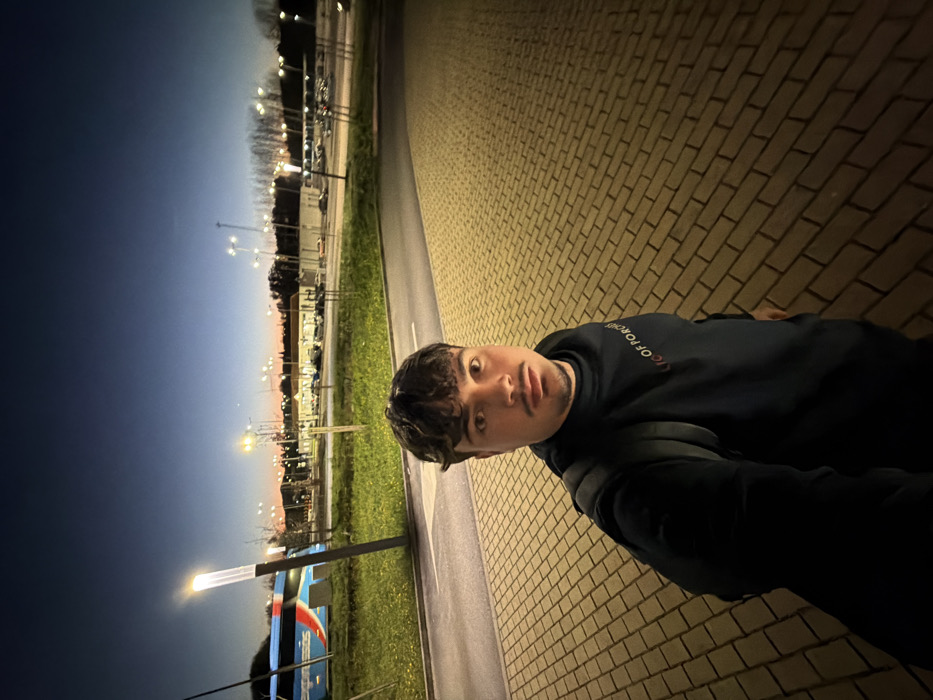 The image depicts a male individual standing on a brick-patterned paved surface in Penafiel, Portugal, during twilight. The sky transitions from dark blue overhead to an orange-pink glow on the horizon. Artificial lighting from numerous lampposts illuminates the scene.
The man, positioned centrally and slightly to the left, is taking a selfie, holding the camera at arm's length. He has short, curly dark brown hair, a light mustache, and a short goatee. He is wearing a dark, possibly black or navy blue, long-sleeved hooded sweatshirt with a white, red, and yellow graphic on the chest, partially visible as "OF POR." He also wears a dark backpack.
Behind the individual, a narrow asphalt road or bus lane runs horizontally. To the left of the road, a grassy median strip features a single, illuminated lamppost with a rectangular light fixture, and some small plants. A large blue and white "EXPRESSOS" branded bus is parked on this side of the road.
Further in the background, a sprawling paved area, likely a parking lot, is visible, containing multiple parked vehicles and illuminated by numerous tall lampposts emitting a yellowish light. Beyond this area, commercial-style buildings and a distant treeline are discernable. The overall environment suggests a bus terminal or a large commercial complex with significant parking infrastructure.