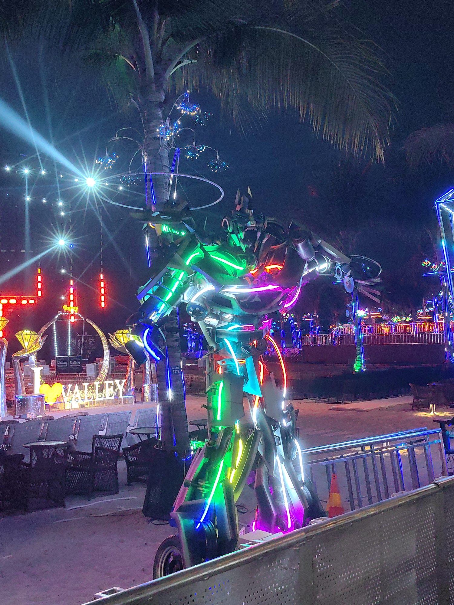 The image depicts an illuminated robot figure at night in what appears to be an entertainment area. The robot is adorned with bright, multicolored LED lights, predominantly green, blue, white, and pink. It stands on wheels and is partially obscured by a palm tree trunk.
In the background, the words "I ❤️ VALLEY" are prominently displayed, indicating a location named "Valley." Further back, there are more illuminated decorations and structures, some resembling diamonds and a crescent moon. The presence of tables and chairs suggests an outdoor seating area. A fence can be seen in the foreground.
The image was taken in Bai Chay Ward, Vietnam.