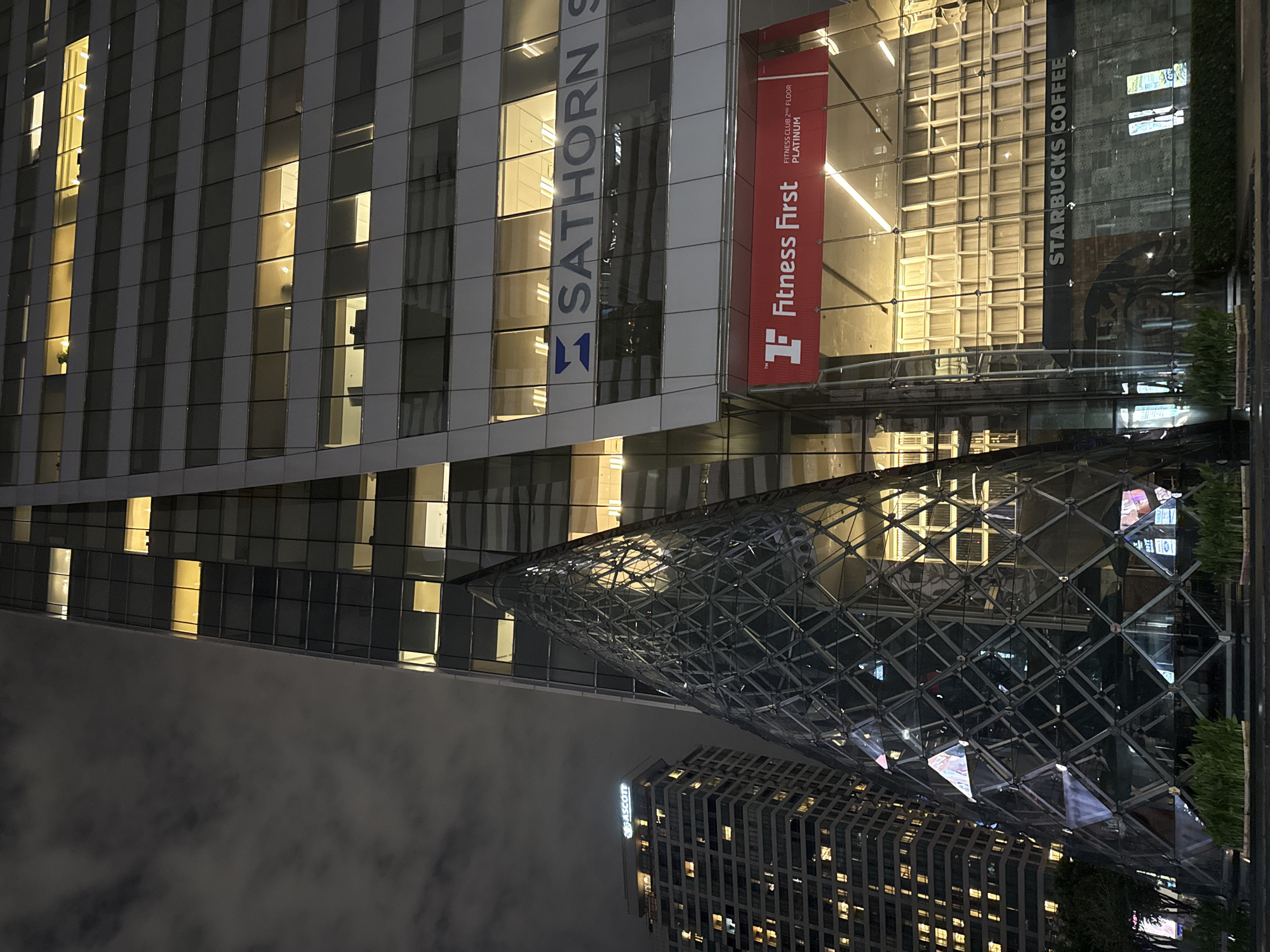 This media file depicts a nighttime scene in a modern urban environment, likely Bangkok, Thailand. The primary subjects are contemporary architectural structures, including a tall office building with a grid of illuminated windows and a unique, faceted glass structure that appears to be an architectural element or a separate building.
The office building features prominent signage: "SATHORN" is visible in large, blue lettering on its facade, and a red banner with white text reads "Fitness First" along with "FITNESS CLUB 22ND FLOOR PLATINUM". Another vertical sign displays "STARBUCKS COFFEE". The windows of the office building are lit, suggesting activity within, though no individuals are clearly discernible. The glass structure reflects the lights of the surrounding buildings and the sky. In the distance, another building with numerous illuminated windows, possibly a hotel or residential complex, is visible. The sky is dark and cloudy, indicating a nighttime setting. The overall mood is one of urban vibrancy and commercial activity, even at night.