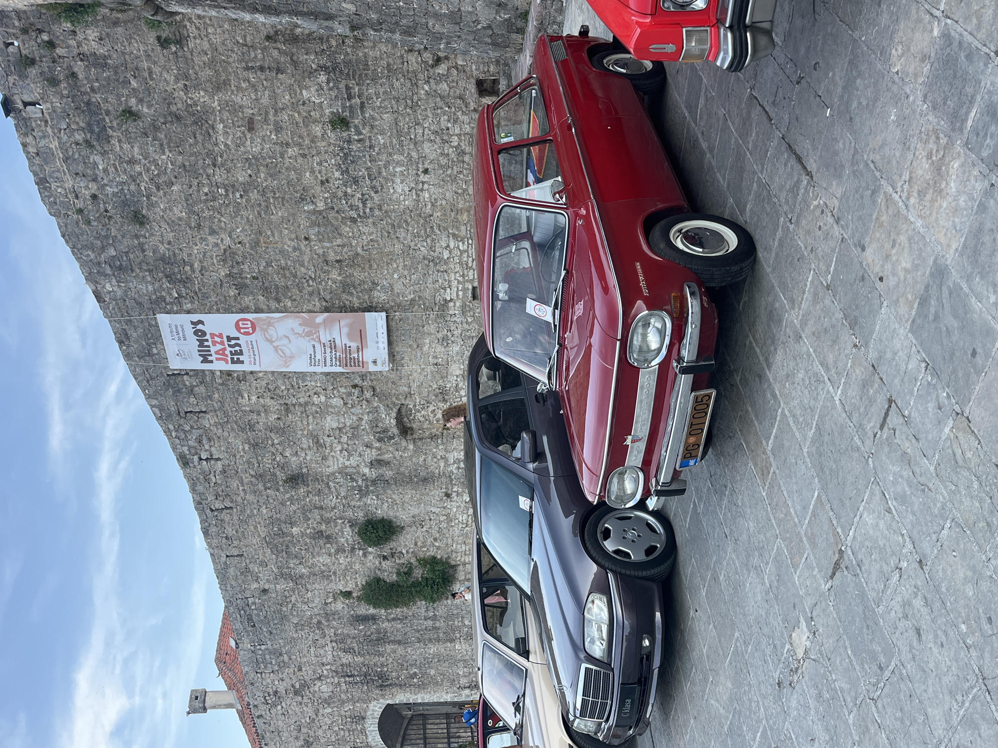 The image shows a street scene in the old town of Budva, Montenegro. Several parked cars are visible, including a red Zastava 1300 "Tristać" with license plate PG OT005, a dark purple Mercedes-Benz C-Class, and another car partially visible. The red Zastava 1300 "Tristać" appears to be a classic car, suggesting an event or gathering.
In the background is a stone wall, possibly part of the old city fortifications. A banner is attached to the wall promoting the "Mimo's Jazz Fest," scheduled for May 20-23, 2026, at the Stari Grad (old town) Budva. The banner lists performers such as Vlatko Stefanovski Trio and MIMO Sextet. There are also some people faintly visible on the background. The overall setting is outdoors on a paved street. The environment suggests a historical location with a blend of cultural events.