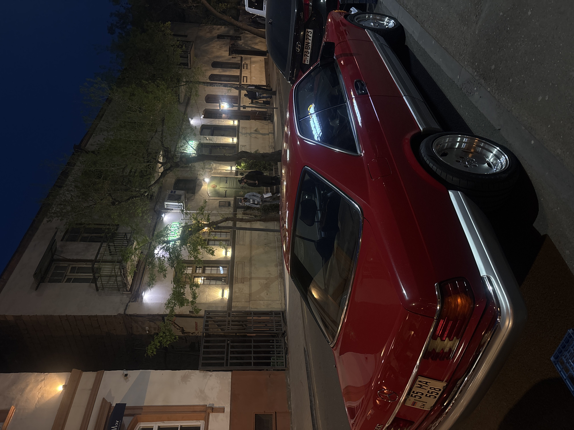 The image shows a red Mercedes-Benz coupe parked on a street in Yerevan, Armenia at night. A dark-colored car is parked behind the Mercedes. Pedestrians are visible on the sidewalk near a building with multiple lighted windows. The street is dimly lit. The license plate of the red car reads "55 MA 558" with the letters "AM" indicating Armenia.