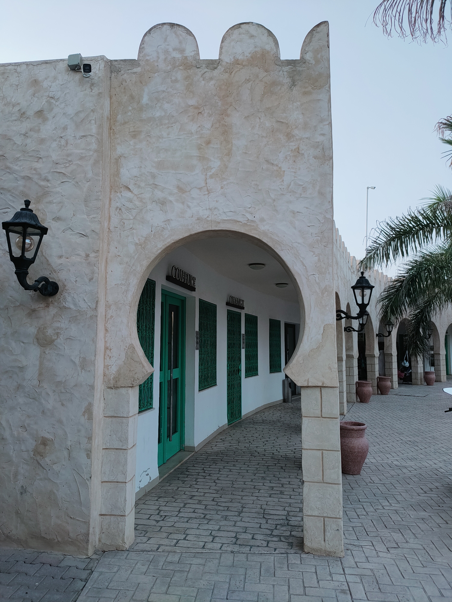 The image shows a picturesque outdoor scene in Nabeul, Tunisia, with a distinctive architectural style. The foreground features a textured, off-white building with a large, arched entryway. The building's facade has a rough, stucco-like finish and is topped with crenellations, reminiscent of historical fortifications. To the left of the archway, a black, wrought-iron lantern with a visible bulb is mounted on the wall.
The arched entryway leads to a walkway with a patterned brick floor. On the left side of the walkway, a green door is visible, flanked by green-shuttered windows. Above the door, a sign reads "COIFFURE" in dark lettering. Further down the walkway, through a series of similar arches, more green doors and windows can be seen, suggesting a row of shops or establishments.
To the right of the main archway, another black lantern is mounted on a pillar. In the background, palm trees with fronds extending into the pale blue sky are visible, hinting at a warm climate. Several large, reddish-brown clay pots are placed along the walkway, adding to the decorative elements of the scene. The overall impression is one of a well-maintained, possibly tourist-oriented area, characterized by traditional North African architectural influences.