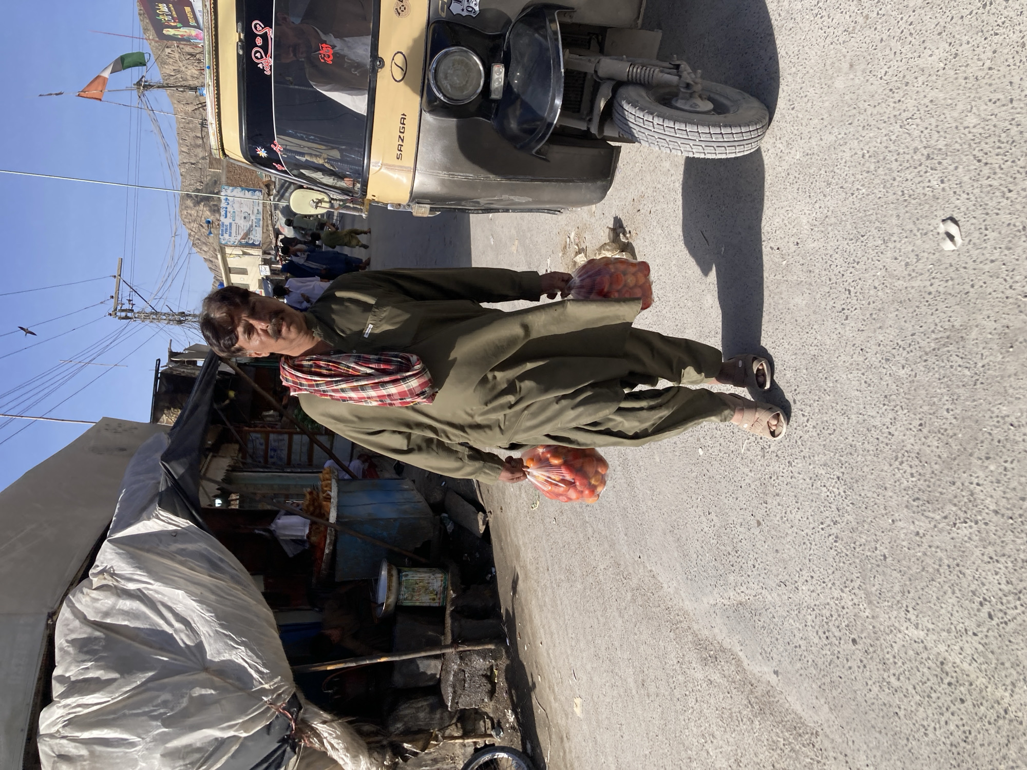 A man in Quetta City Tehsil, Pakistan, stands on a paved street next to a three-wheeled auto rickshaw vehicle. He is dressed in a long-sleeved green outfit with a red and white checkered scarf around his neck and sandals on his feet. He holds two transparent plastic bags filled with red vegetables, likely tomatoes. The street features a variety of shops and stalls under a large white and black tarp, with various wares displayed. In the background, other pedestrians walk on the street, and mountains are visible in the distance under a clear blue sky.