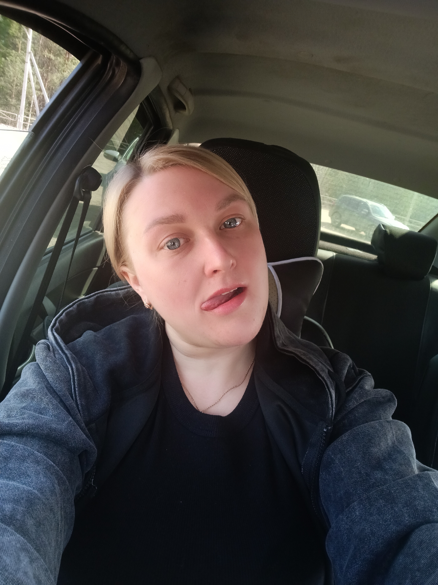 A close-up selfie shows a blonde woman sitting in the driver's seat of a car during the daytime. She looks directly at the camera with a playful expression, her tongue slightly extended. She has blue eyes and a fair complexion. She is wearing a dark, possibly black, long-sleeved shirt layered under a dark grey or faded black hooded jacket. A thin necklace with a small pendant and small hoop earrings are visible.
The car's interior is visible around her, including parts of black headrests, the door frame, seatbelts, and the ceiling. Through the car windows, an outdoor scene unfolds under bright, sunny weather. Through the front side window, trees and some white structural elements, possibly pylons or fences, are visible. Through the rear window, another dark-colored car, likely an SUV, is partially visible in the distance, suggesting a road or parking area. The lighting indicates a clear and bright day. The overall mood of the image is casual and lighthearted. There is no visible text in the image. The location context for this image is Chaikovsky, Russia.